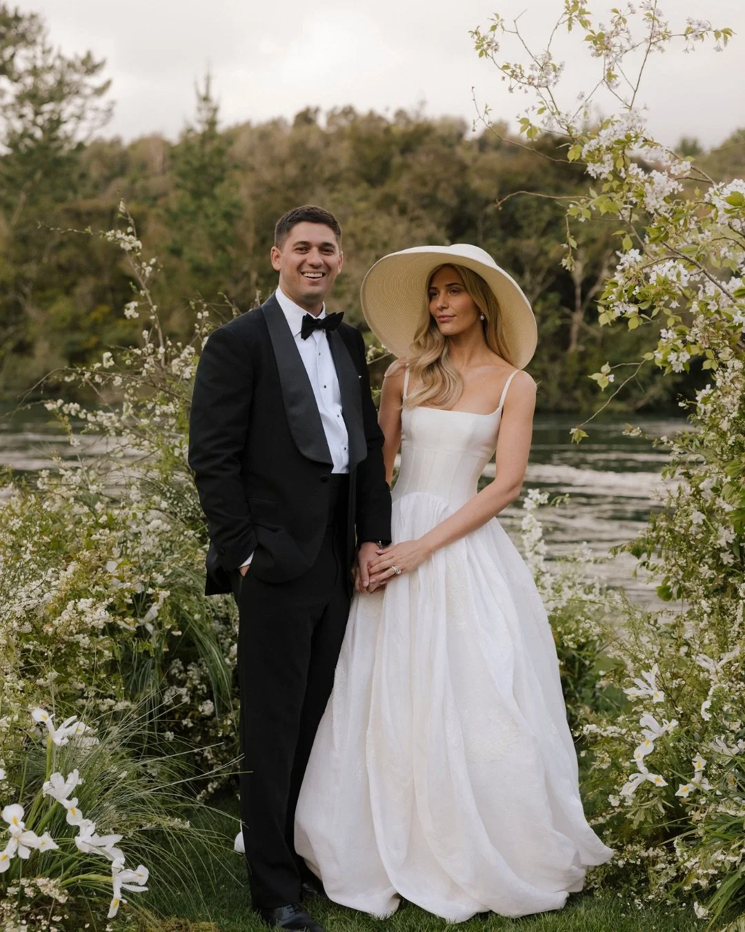 Bride and groom standing outdoors near a river, holding hands, with blooming flowers and trees in the background.