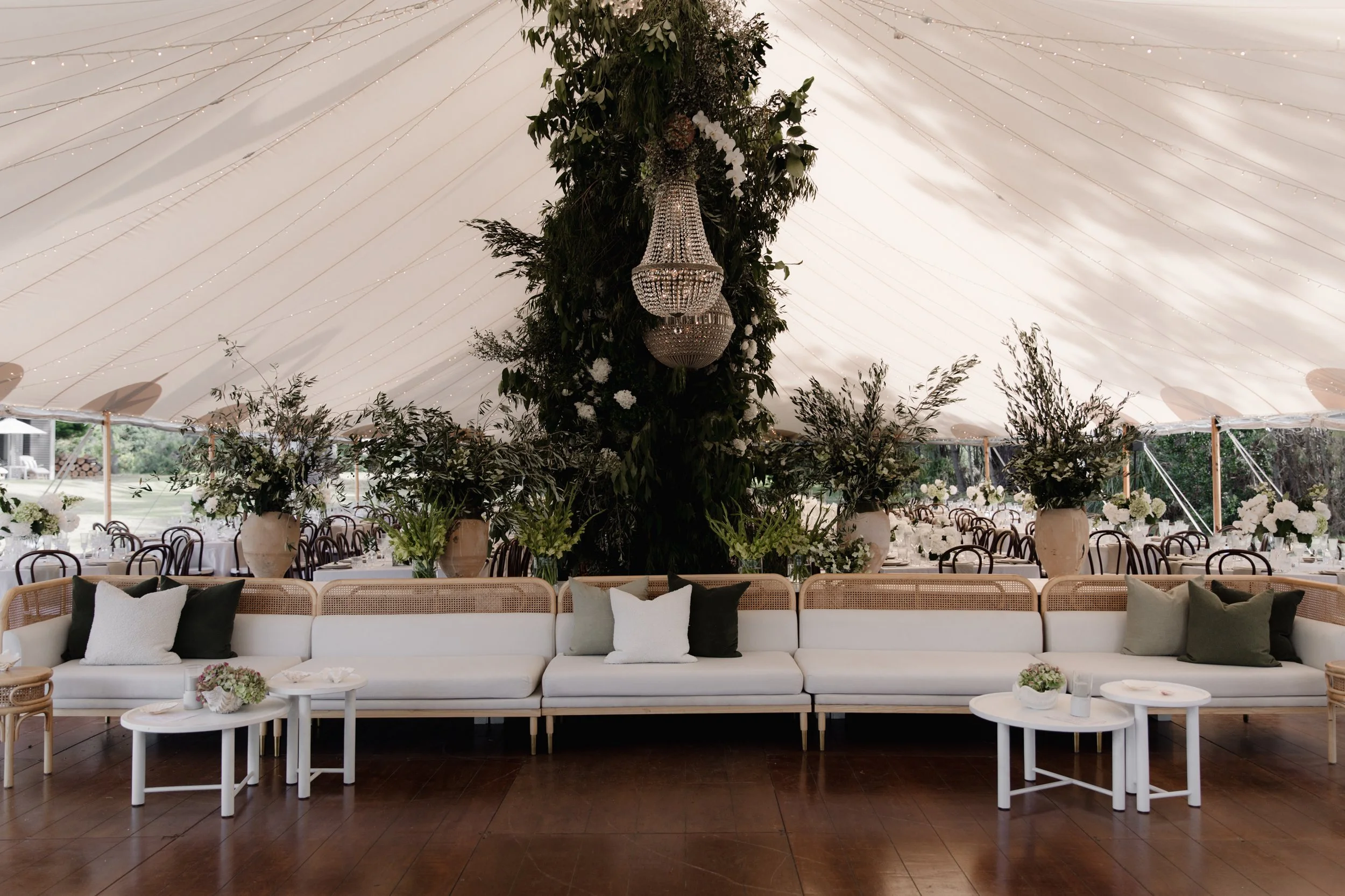 Elegant indoor event decorated with a large floral centerpiece on a draped tent ceiling, surrounded by tables with floral arrangements and seating.