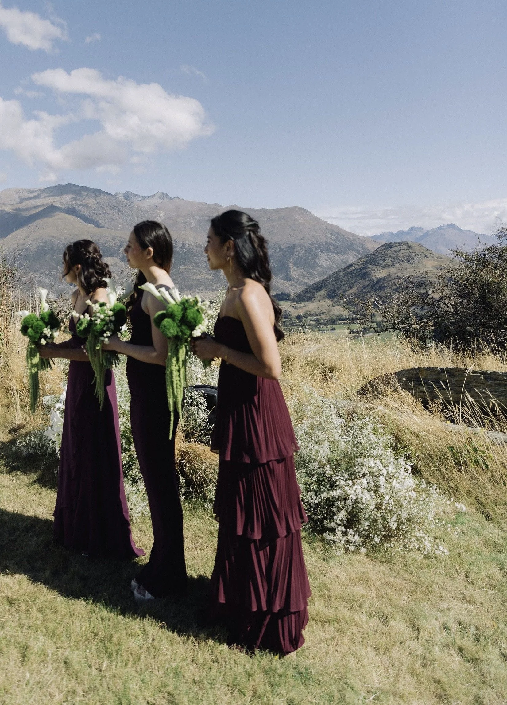 A group of women in formal dresses holding bouquets walk outdoors on a grassy field with mountains and blue sky in the background.