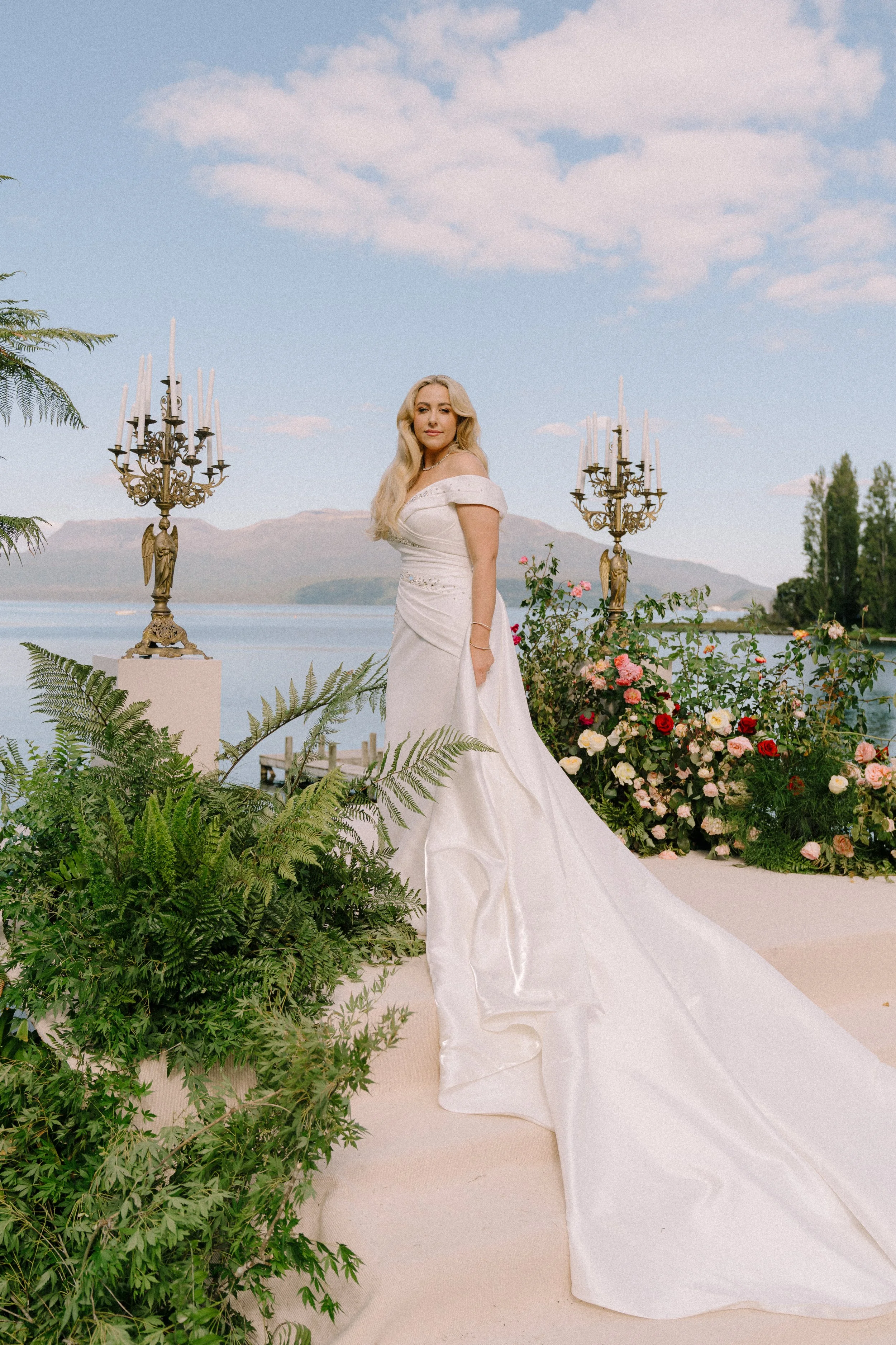A woman in a white, off-the-shoulder wedding dress standing outdoors near a lake, surrounded by pink and red roses and green foliage, with two ornate candelabras behind her and mountains in the distance under a partly cloudy sky.