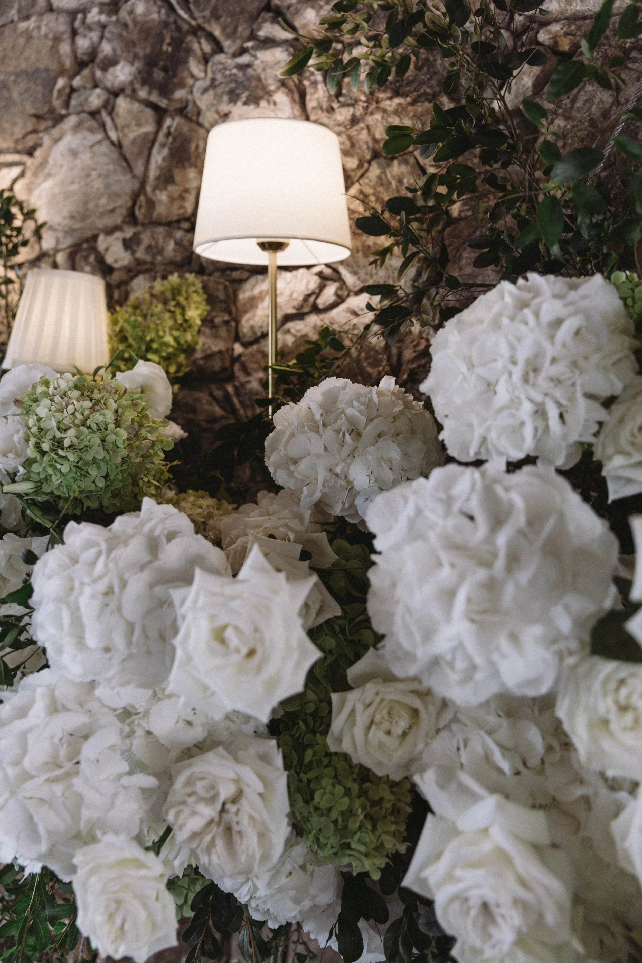 Interior scene with a stone wall, white lampshades, and white hydrangea flowers with green leaves.