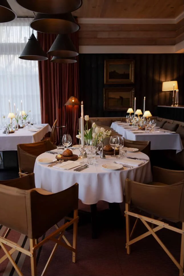 Elegant dining room with round table set for a meal, decorated with white tablecloths, glassware, and candles, surrounded by wooden chairs, with large windows, framed artwork, and warm lighting.