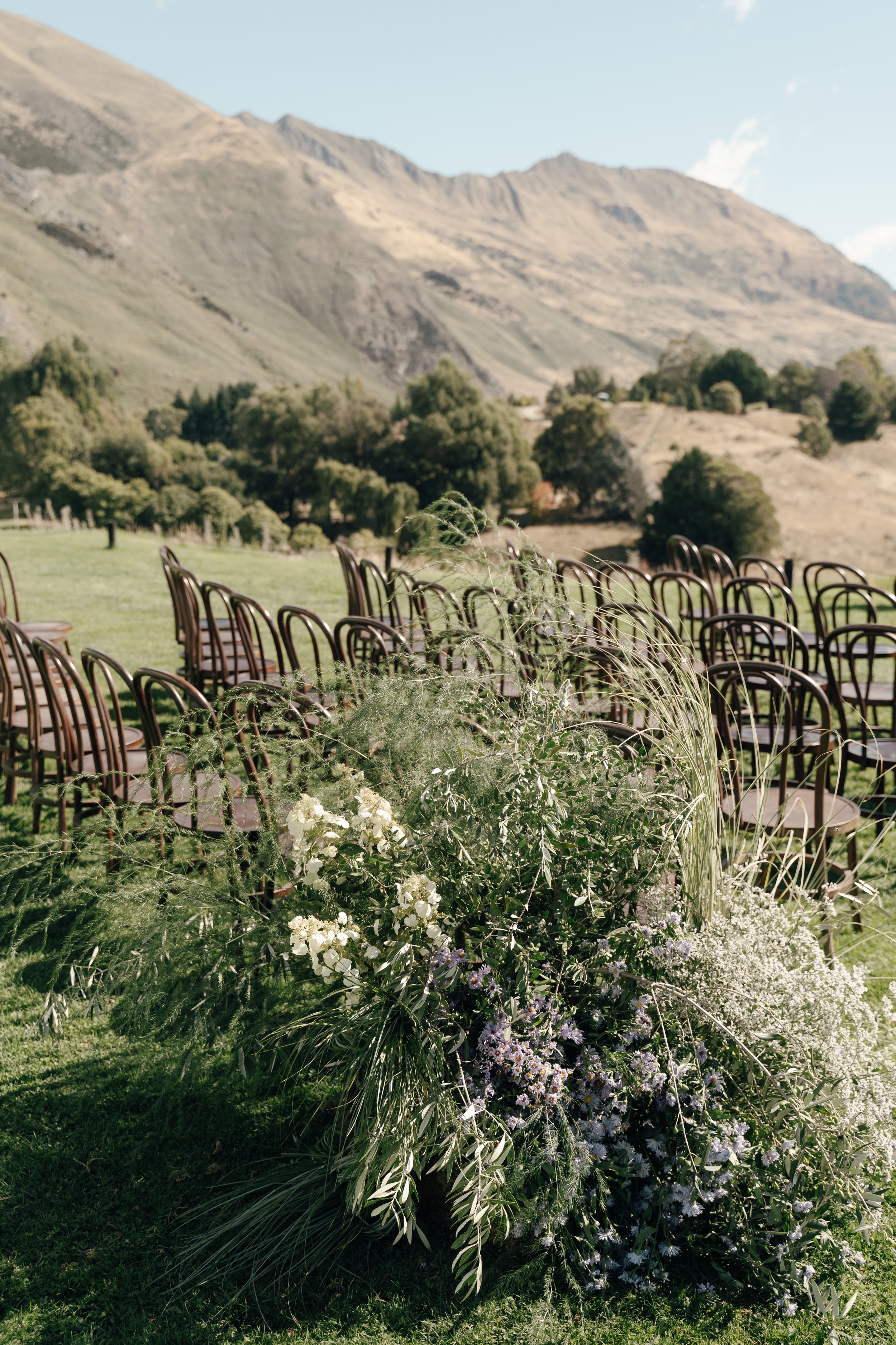 Rows of chairs set up outdoors on a grassy area with a mountain landscape in the background, decorated with arrangements of greenery and white flowers.