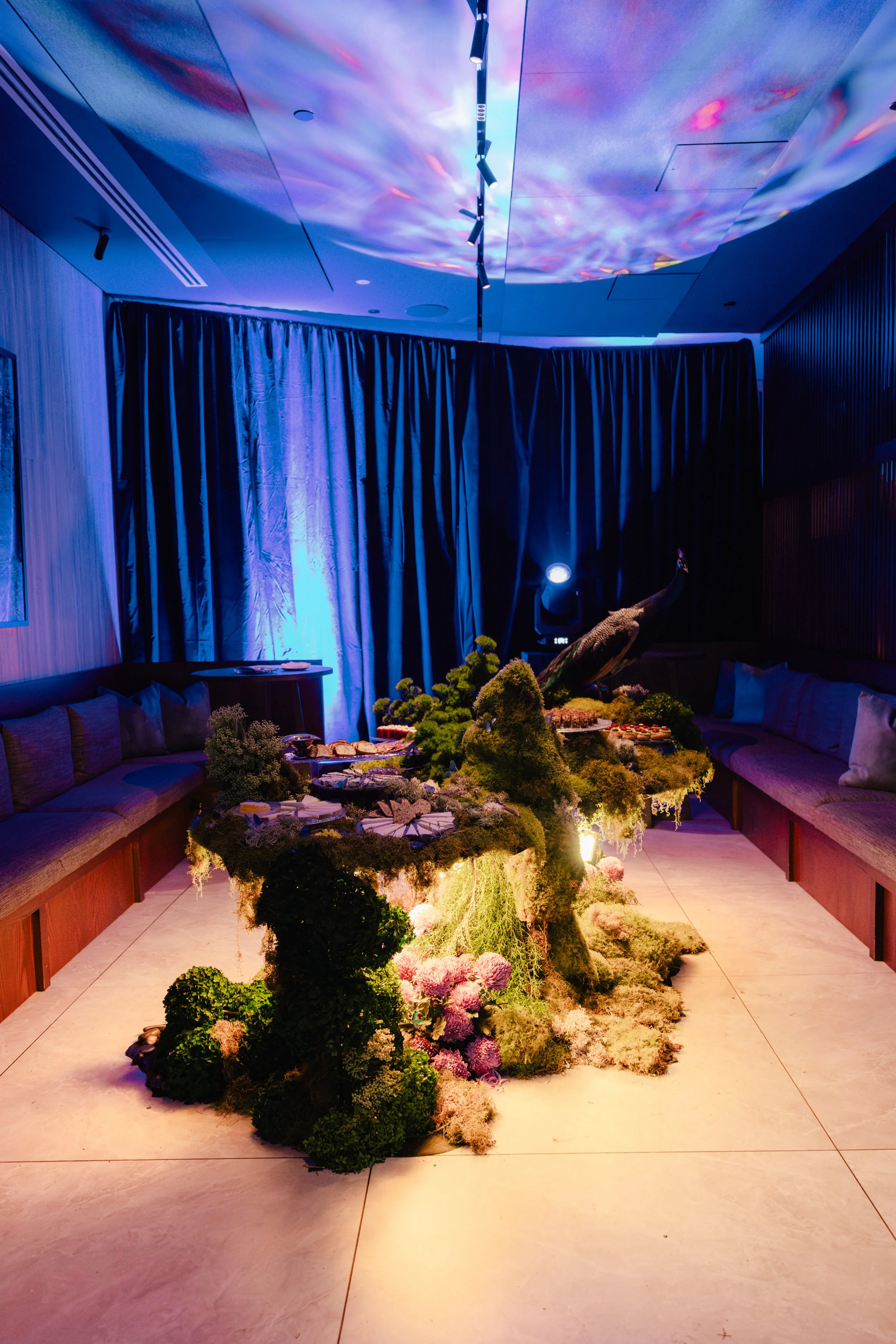 A room with a table decorated with flowers and greenery in the center, surrounded by velvet curtains and cozy seating, illuminated with soft blue and purple lighting, creating a serene atmosphere.