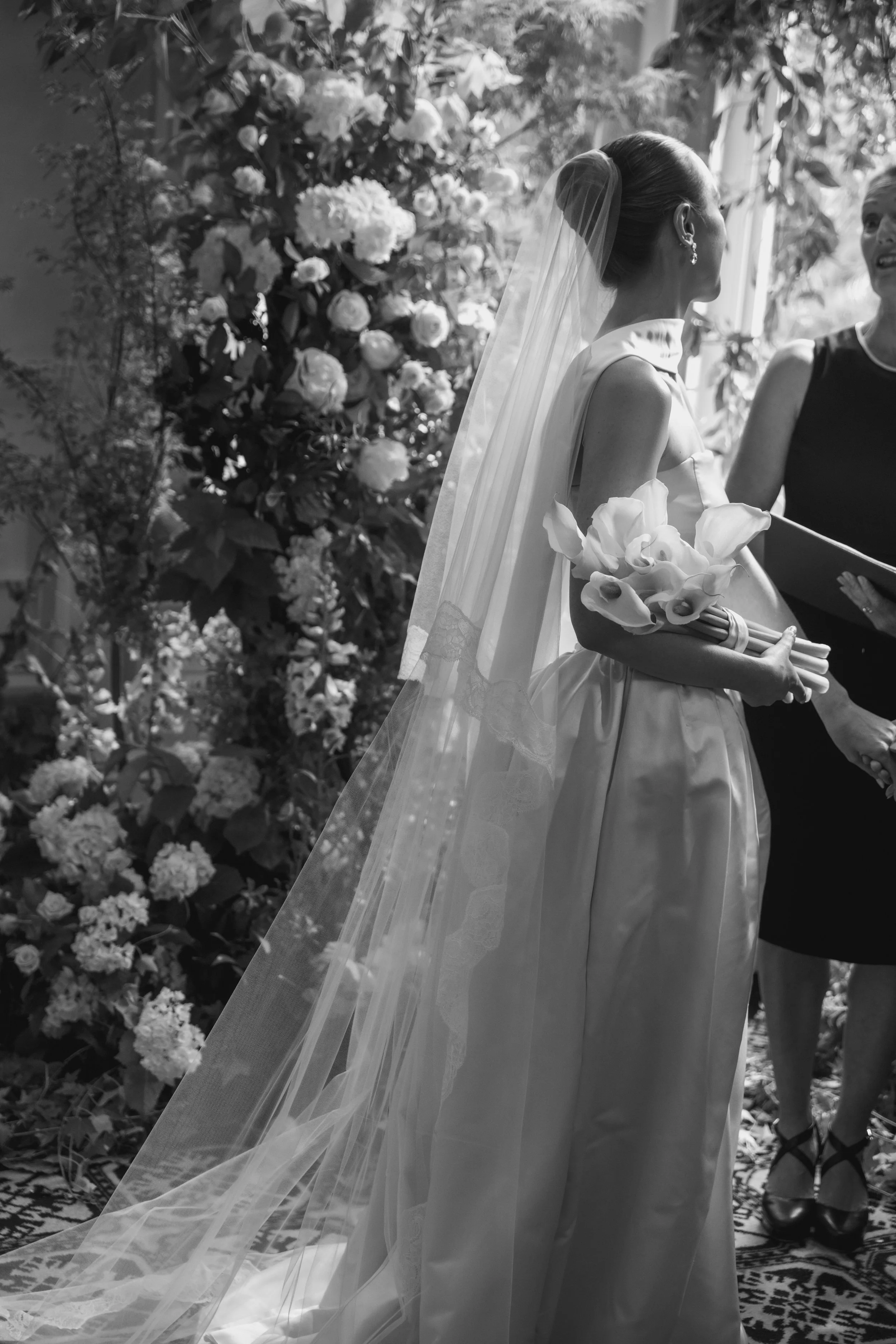 A bride in a wedding gown and veil holding a bouquet of calla lilies, standing near another woman, with floral arrangements in the background, in a black and white photo.
