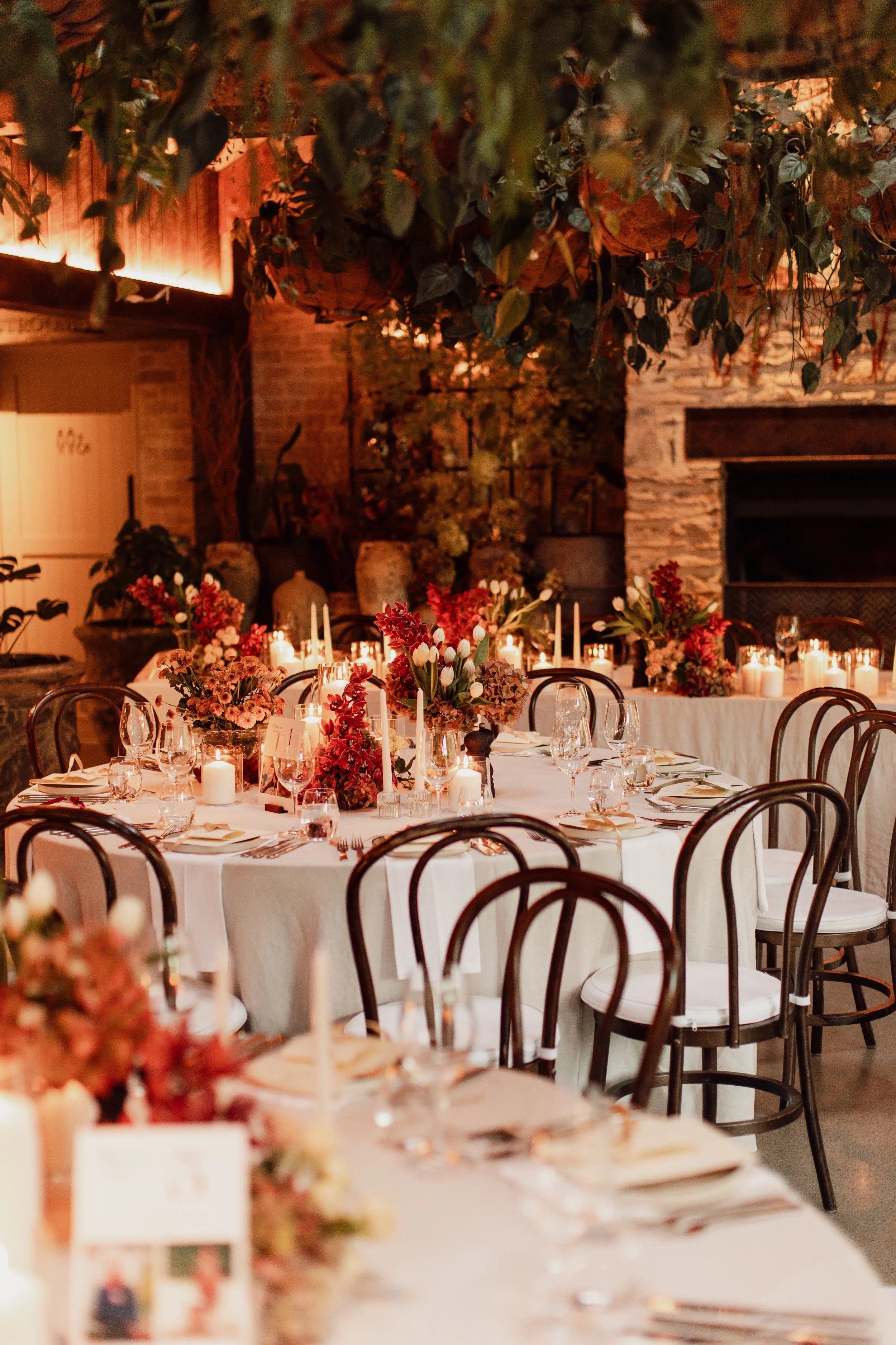 A beautifully decorated indoor dining space with tables set for a formal event, featuring floral centerpieces, candles, and glassware. The room has warm lighting, brick walls, and hanging greenery.