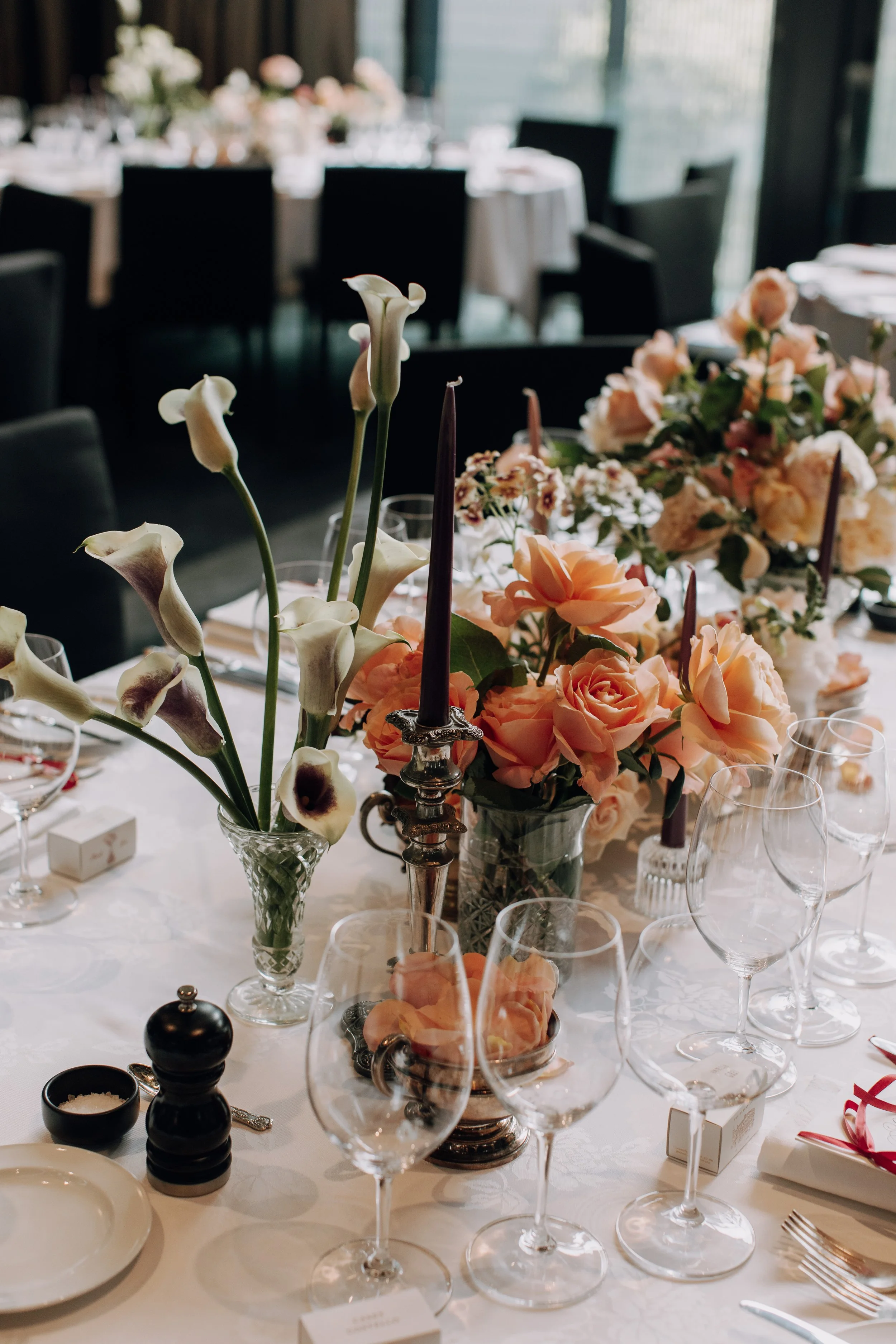 A banquet table decorated with pink and white flowers, tall candlesticks, and multiple wine glasses, set for an elegant event in a room with large windows.
