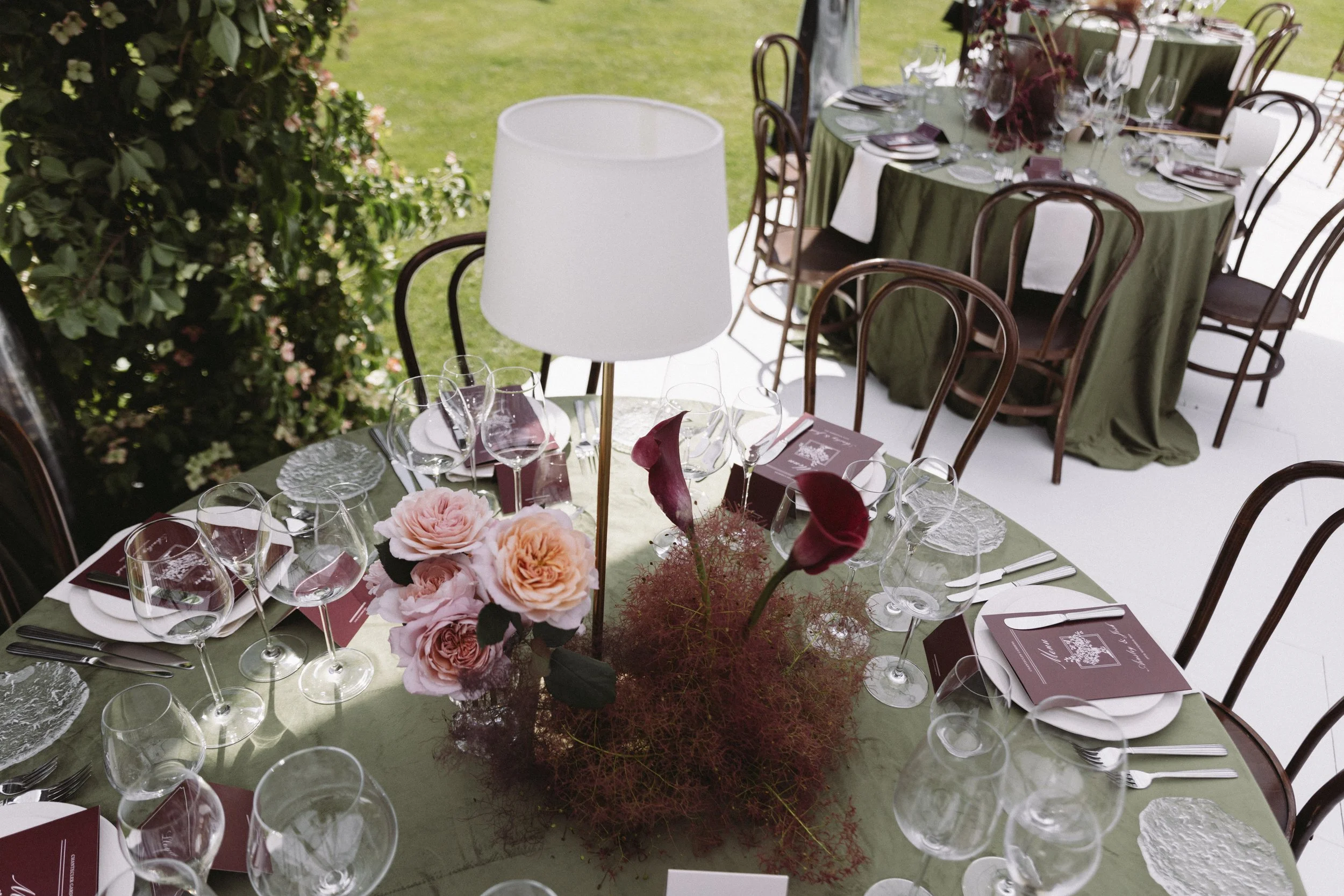 A formal outdoor dining setup with a table decorated with pink flowers, wine glasses, and green tablecloths, surrounded by wooden chairs, on a grassy area.