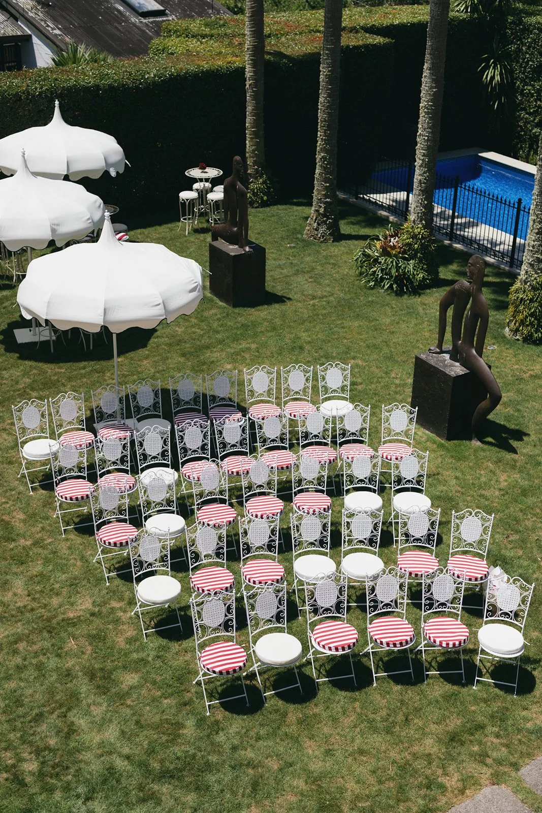 An outdoor area set up for a gathering with white wrought iron chairs with red and white striped cushions arranged in semi-circle, three large white umbrellas providing shade, and decorative statues on black pedestals. There are palm trees, lush green hedges, and a small swimming pool enclosed by a black fence in the background.