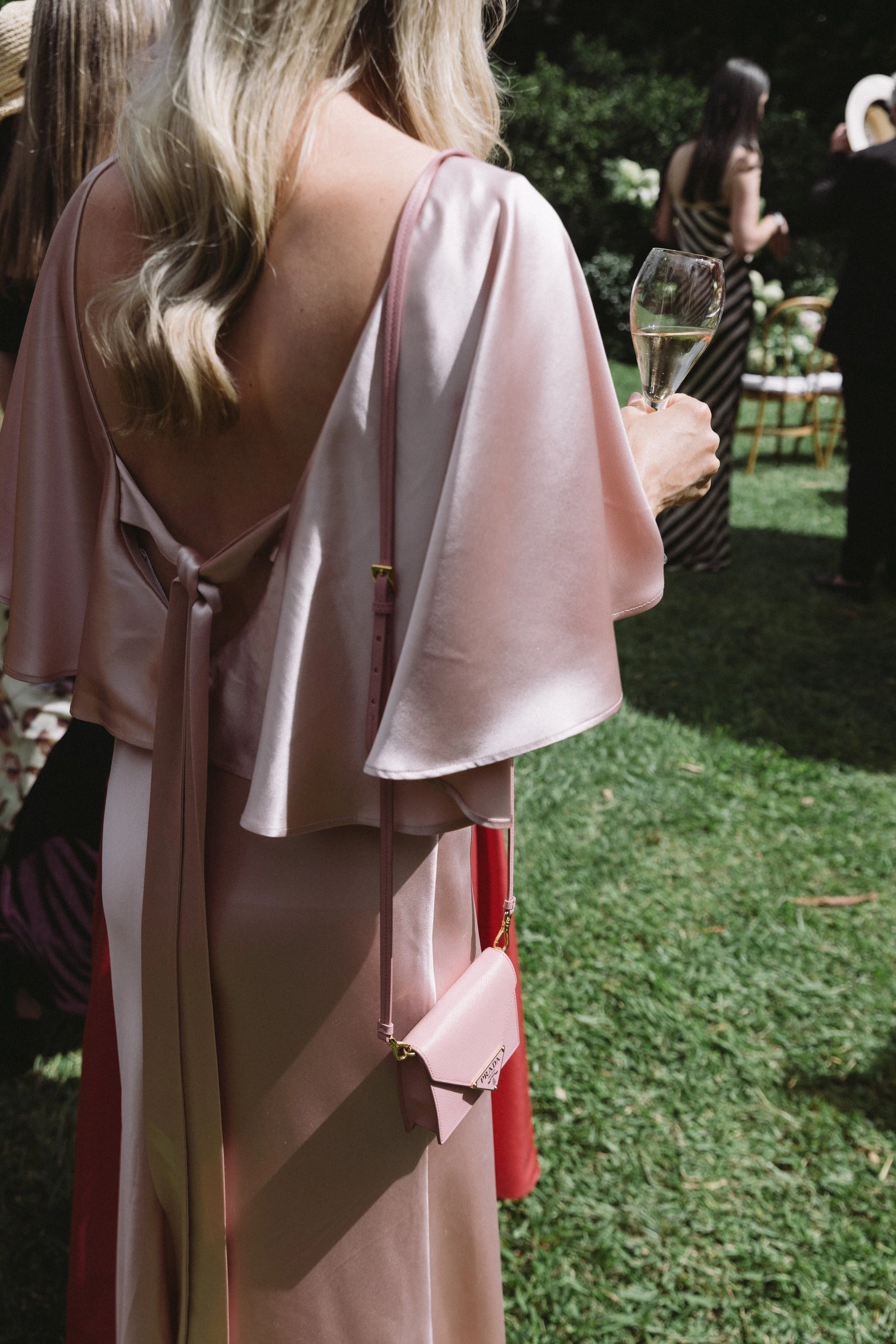 A woman wearing a pink satin cape at an outdoor event, holding a glass of champagne, with other guests and greenery in the background.