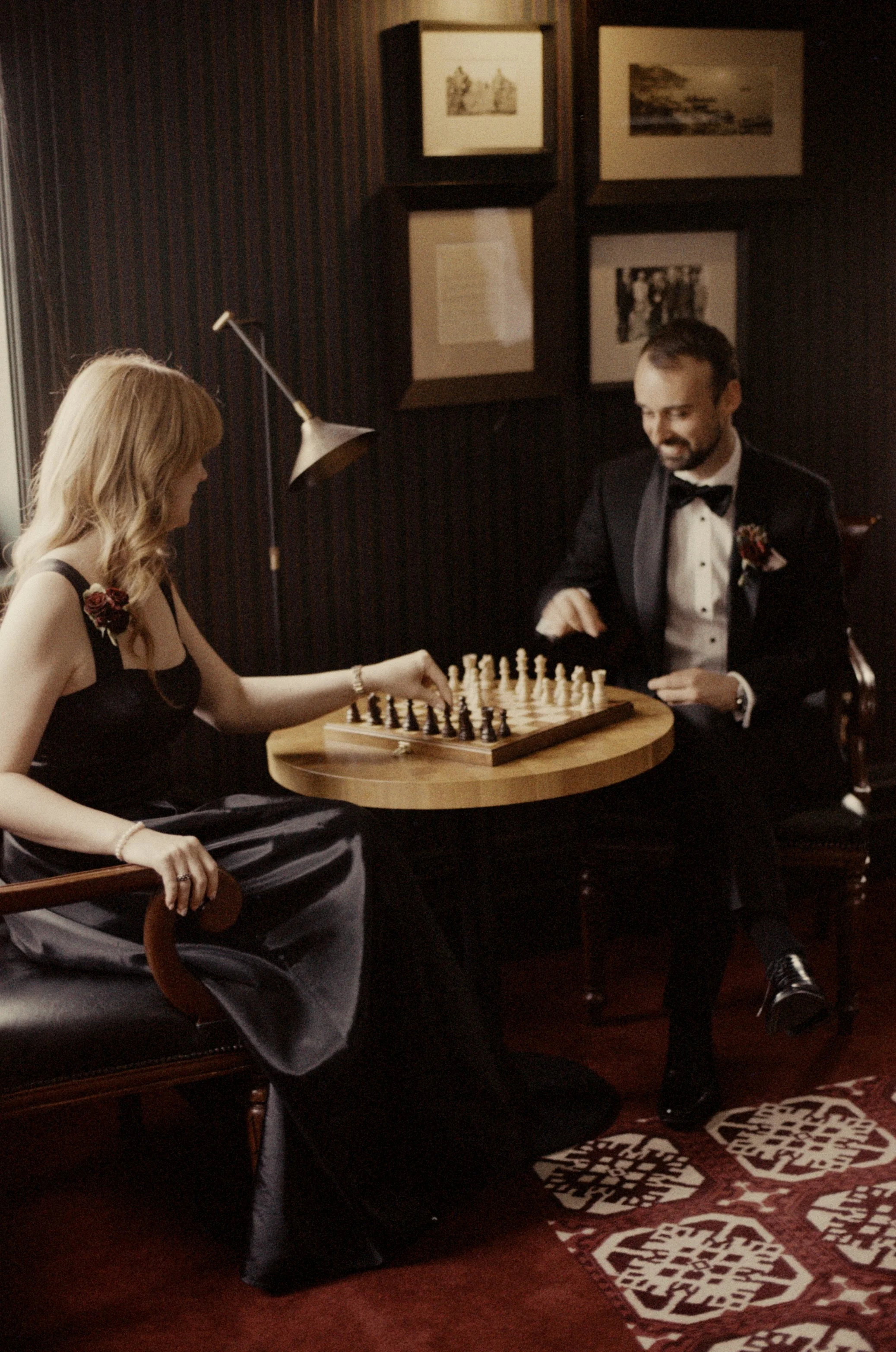 A man and woman playing chess at a table in a dimly lit room, with framed pictures on the dark wall behind them, and a patterned rug on the floor.