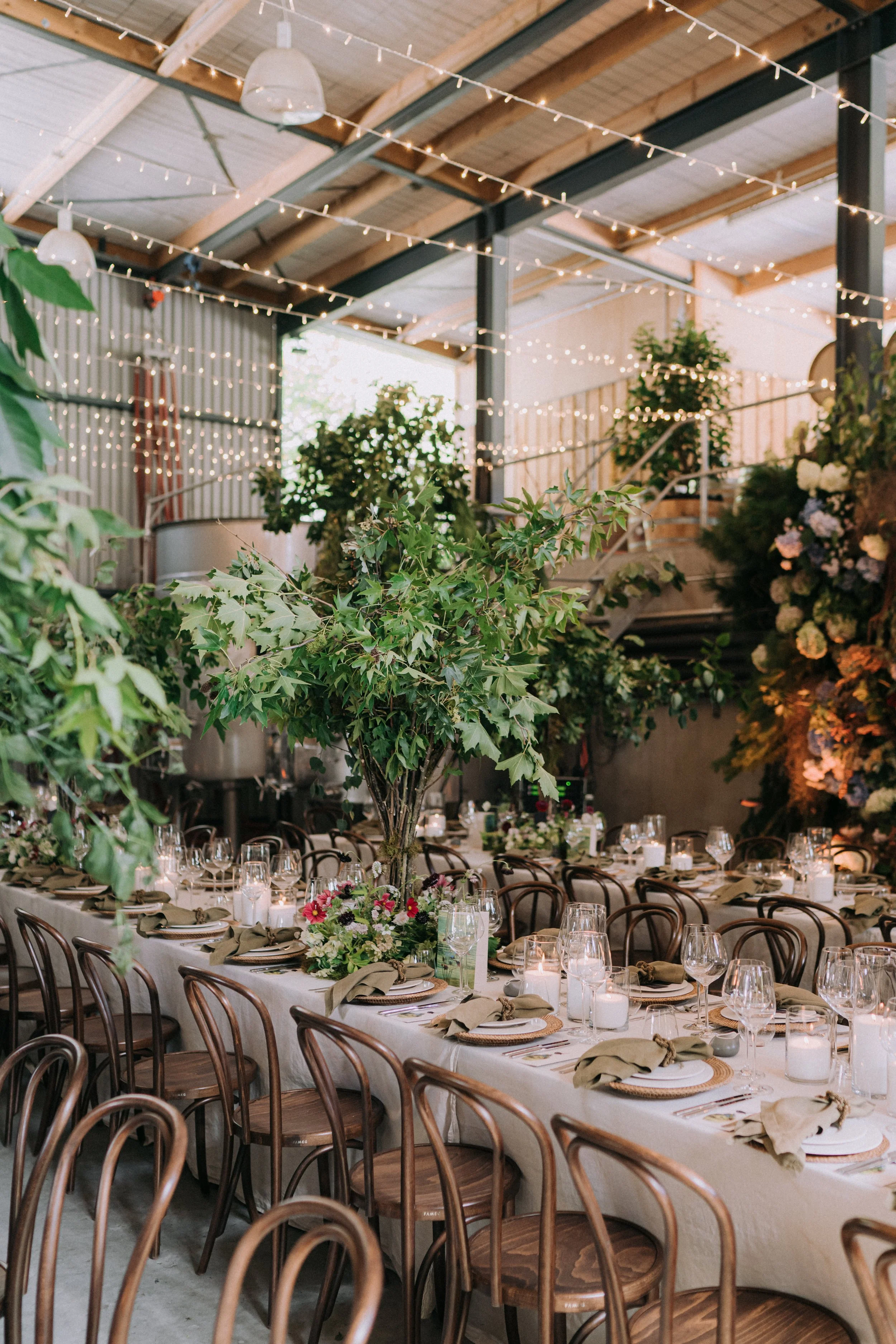 A decorated indoor event space with string lights hanging from the ceiling, plants, and a long banquet table set with plates, glasses, candles, napkins, and floral arrangements for a celebration or wedding reception.