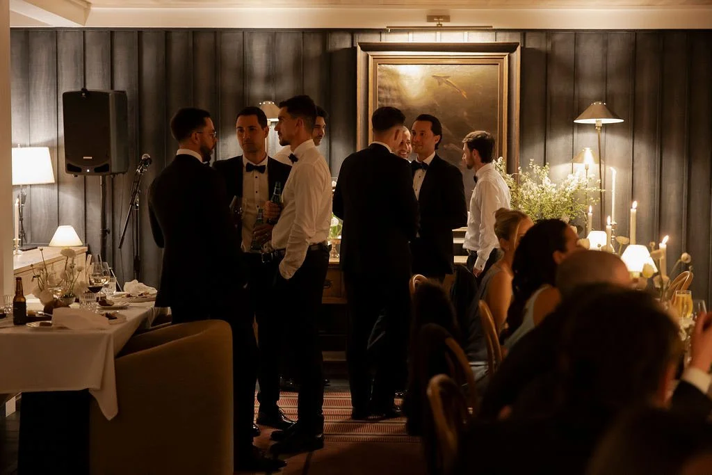Group of men in tuxedos and women in formal attire, socializing and conversing at a dinner party in a warmly lit room with dark wood walls, lamps, and candles.