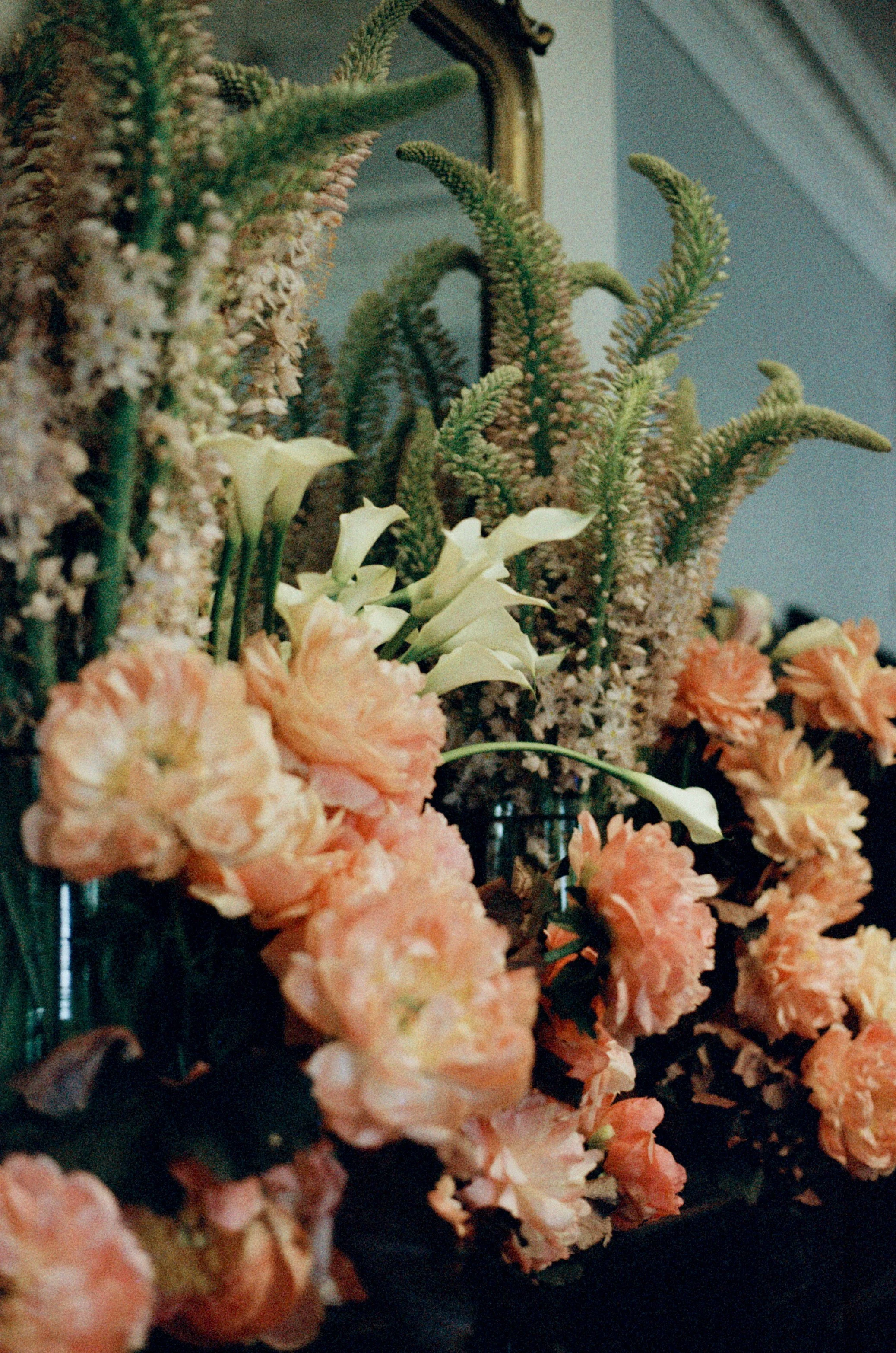 A floral arrangement with light orange carnations, white calla lilies, and green sprigs of pine or fir, set against a dim background with a mirror and a curtain.