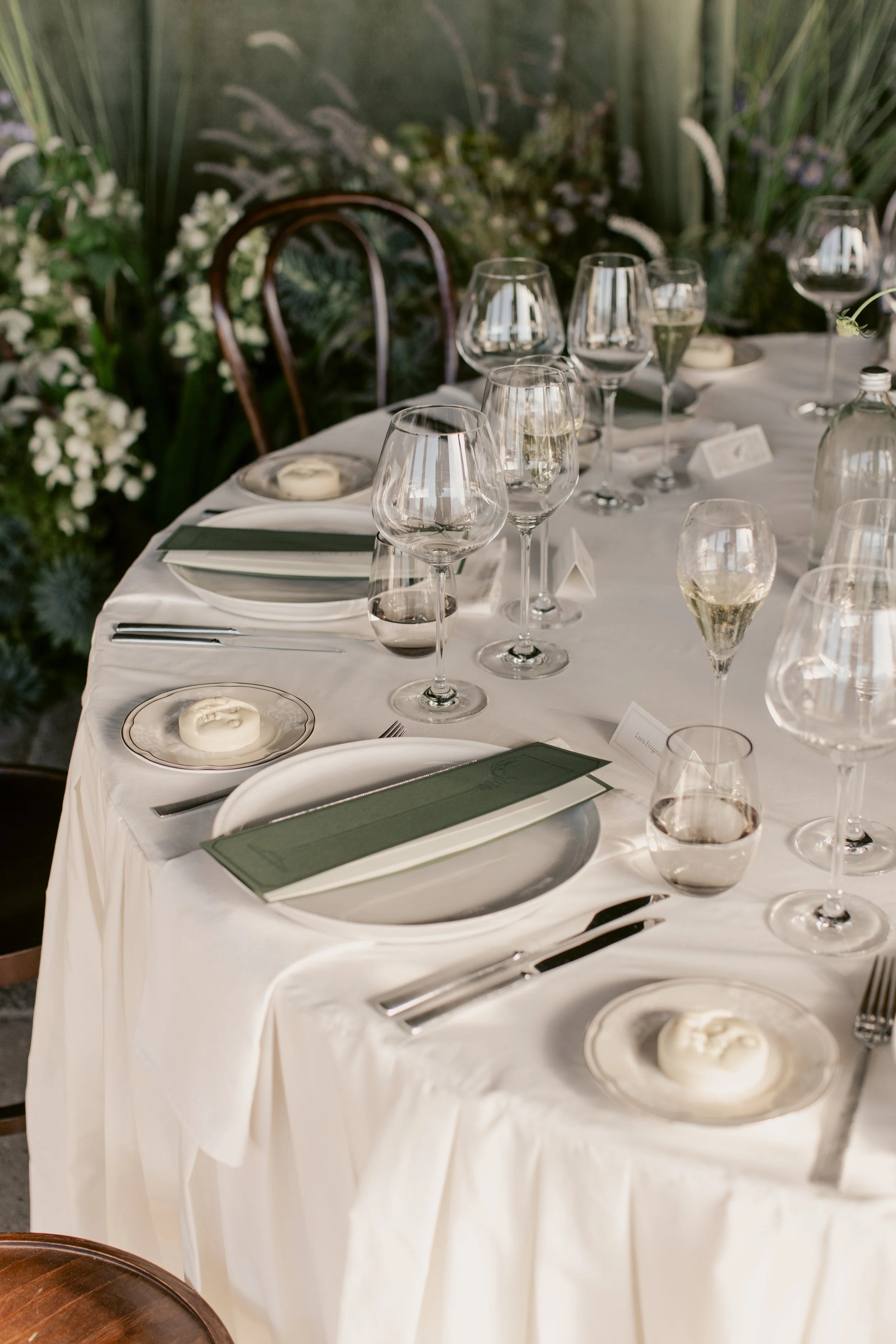 A formal dining table set with wine glasses, plates, napkins, and butter dishes, arranged in a decorated setting with greenery and white flowers in the background.
