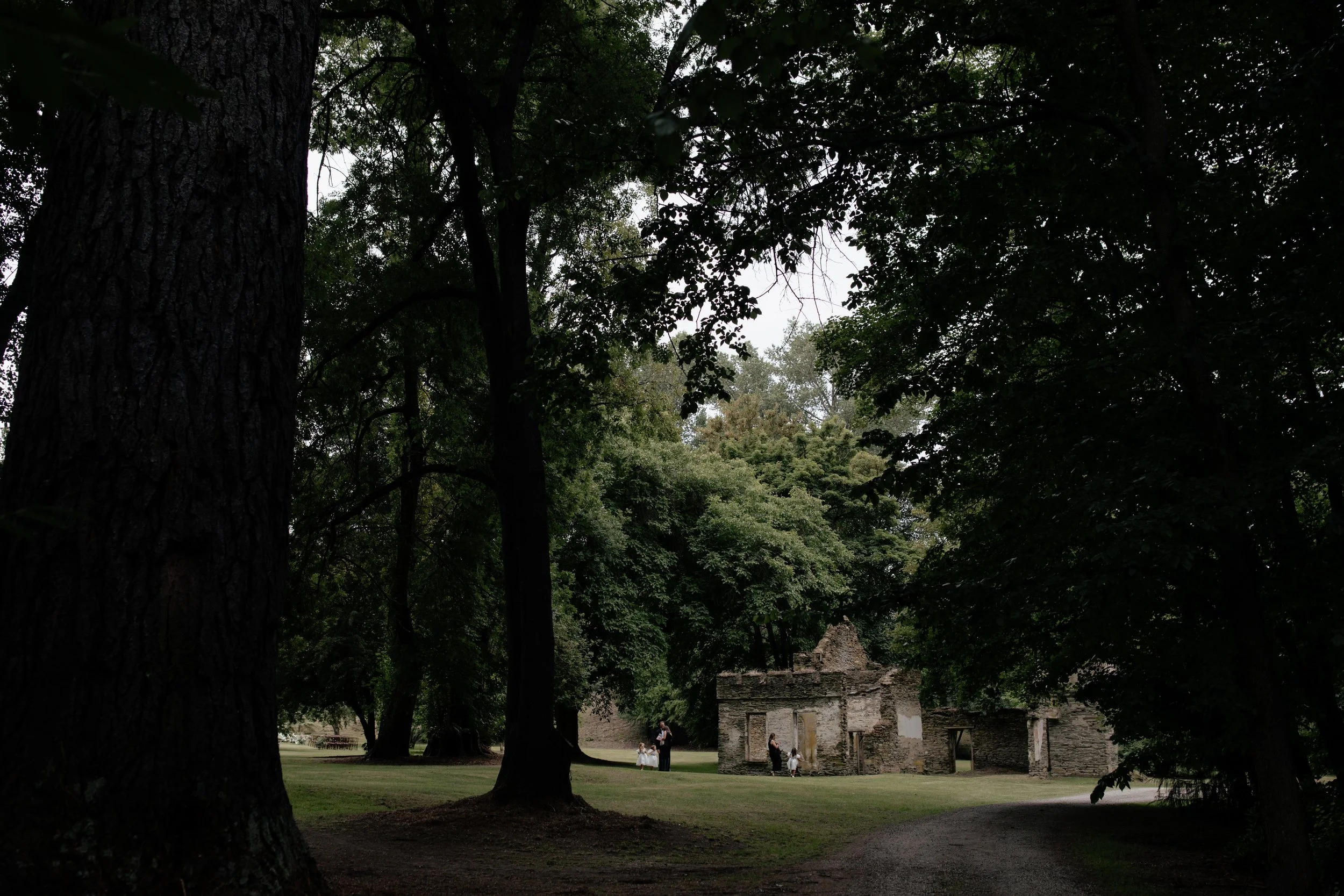 People standing in front of an old stone building with trees overhead in a park, with a green lawn surrounding the structure.