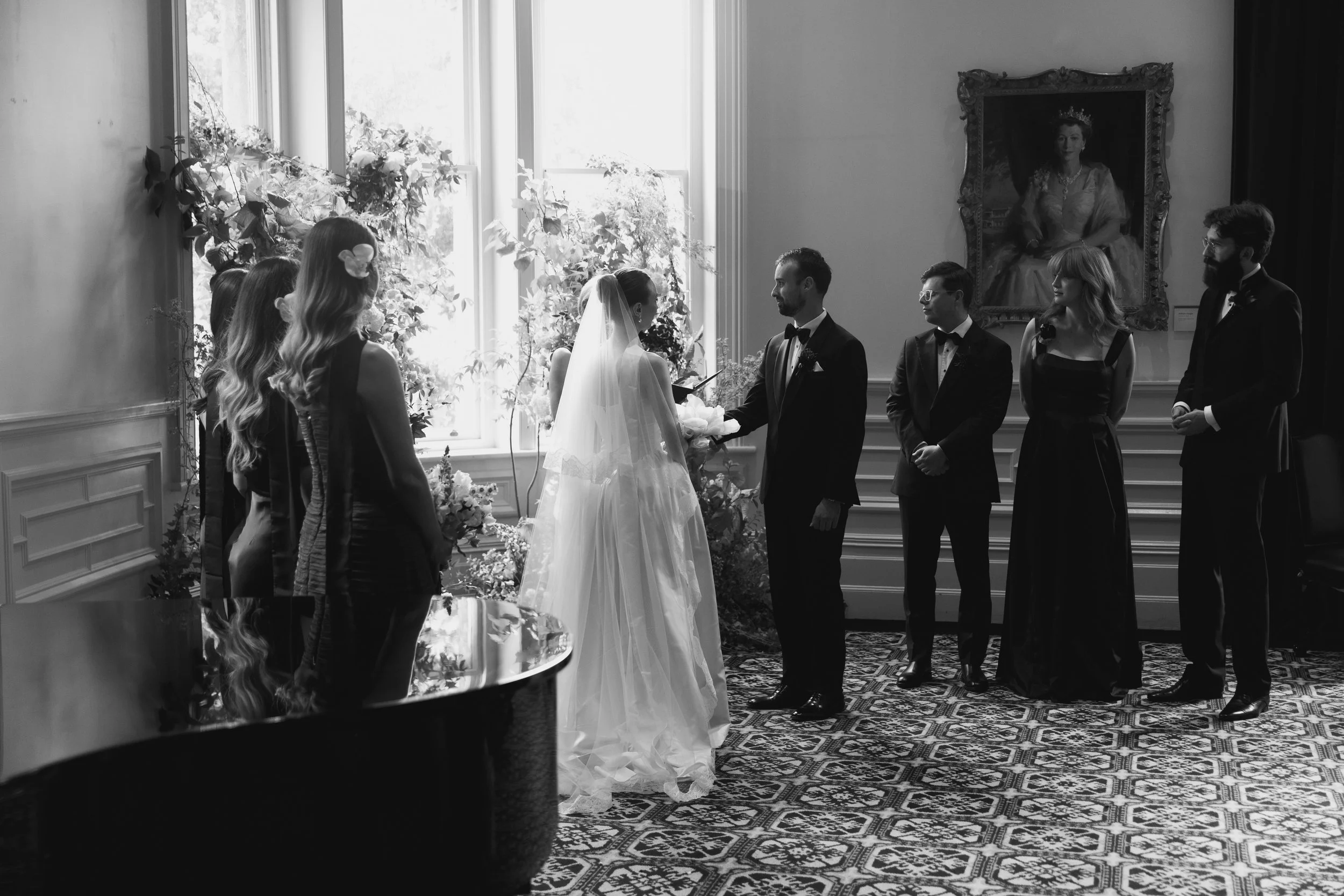 Black and white photo of a wedding ceremony with the bride and groom holding hands in front of their wedding party. The bride is wearing a long gown with a veil, and the groom is in a tuxedo. Wedding guests watch the ceremony. There is a large portrait of a woman in a fancy dress hanging on the wall behind them, and a large window with natural light shines in, illuminating the floral decorations.