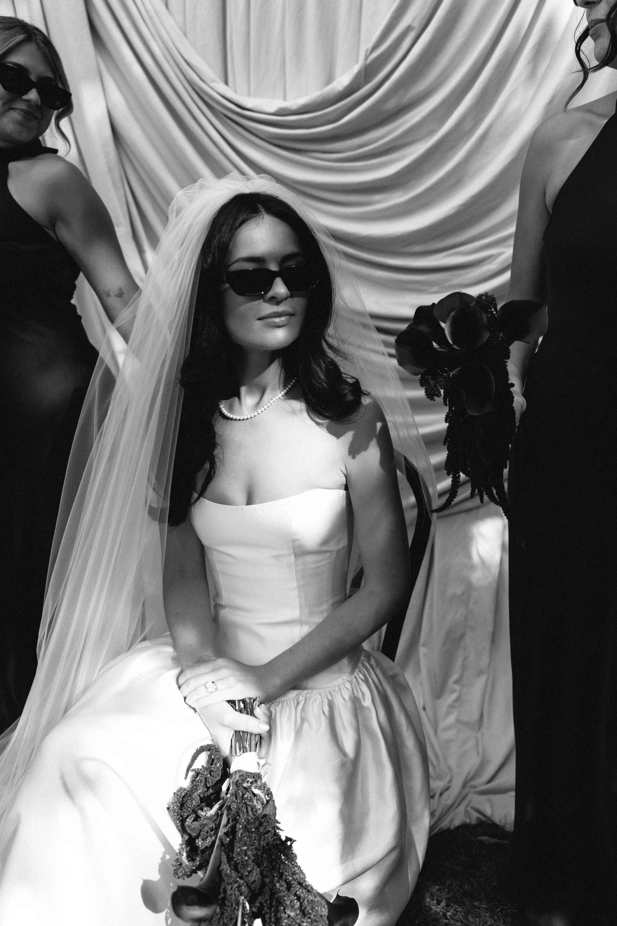 Black and white photo of a woman in a wedding dress with long dark hair, wearing sunglasses and a pearl necklace, sitting while holding a bouquet of flowers. Two women stand beside her, one on each side, dressed in dark outfits, with a draped curtain background.