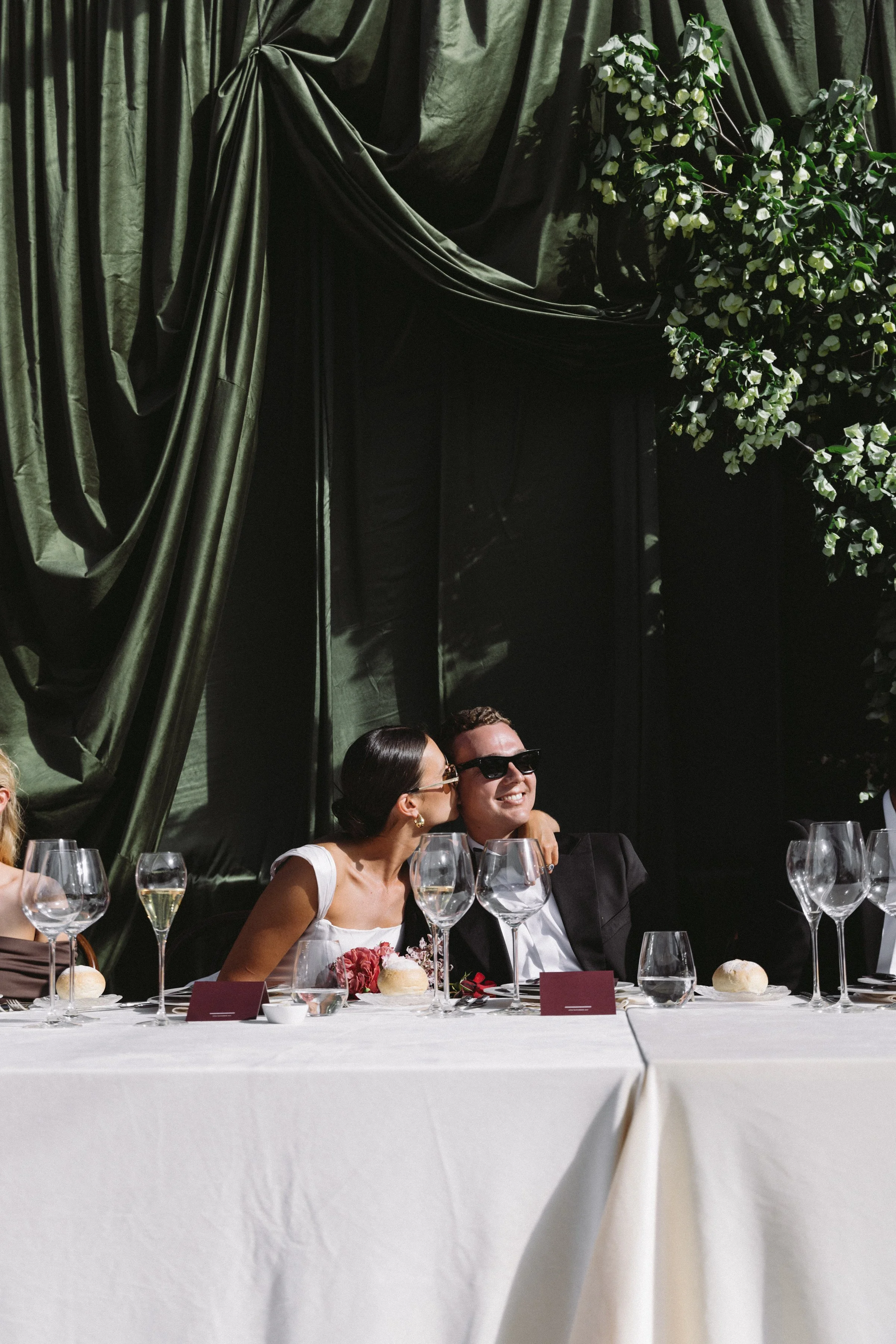 A wedding reception table with two smiling people sitting close to each other, a woman and a man. The table is set with wine glasses, a floral centerpiece, bread rolls, and place cards. There are multiple wine glasses and tableware arranged neatly. The background features dark drapes, green foliage, and a group of people with one woman wearing sunglasses.