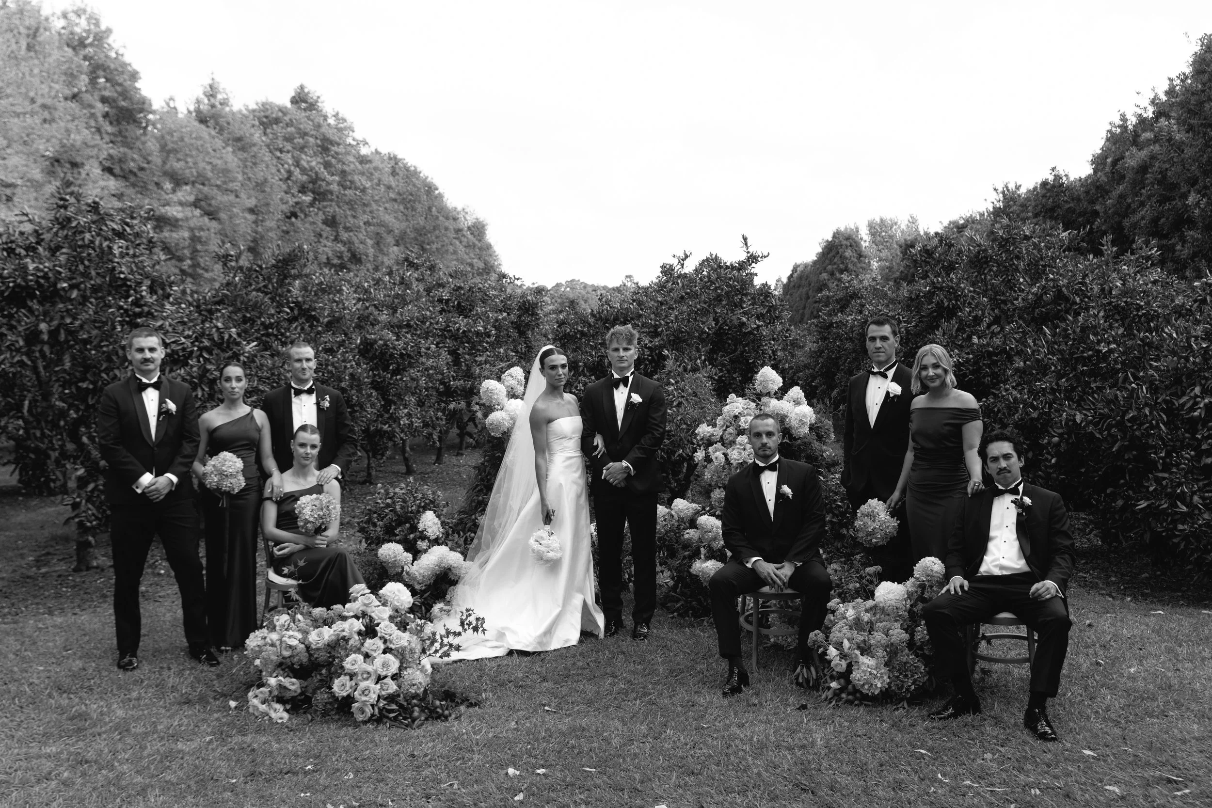 Black and white photo of a wedding party in an outdoor garden with lush trees in the background. The bride and groom are in the center, surrounded by bridesmaids and groomsmen, some standing and some seated, with floral decorations.