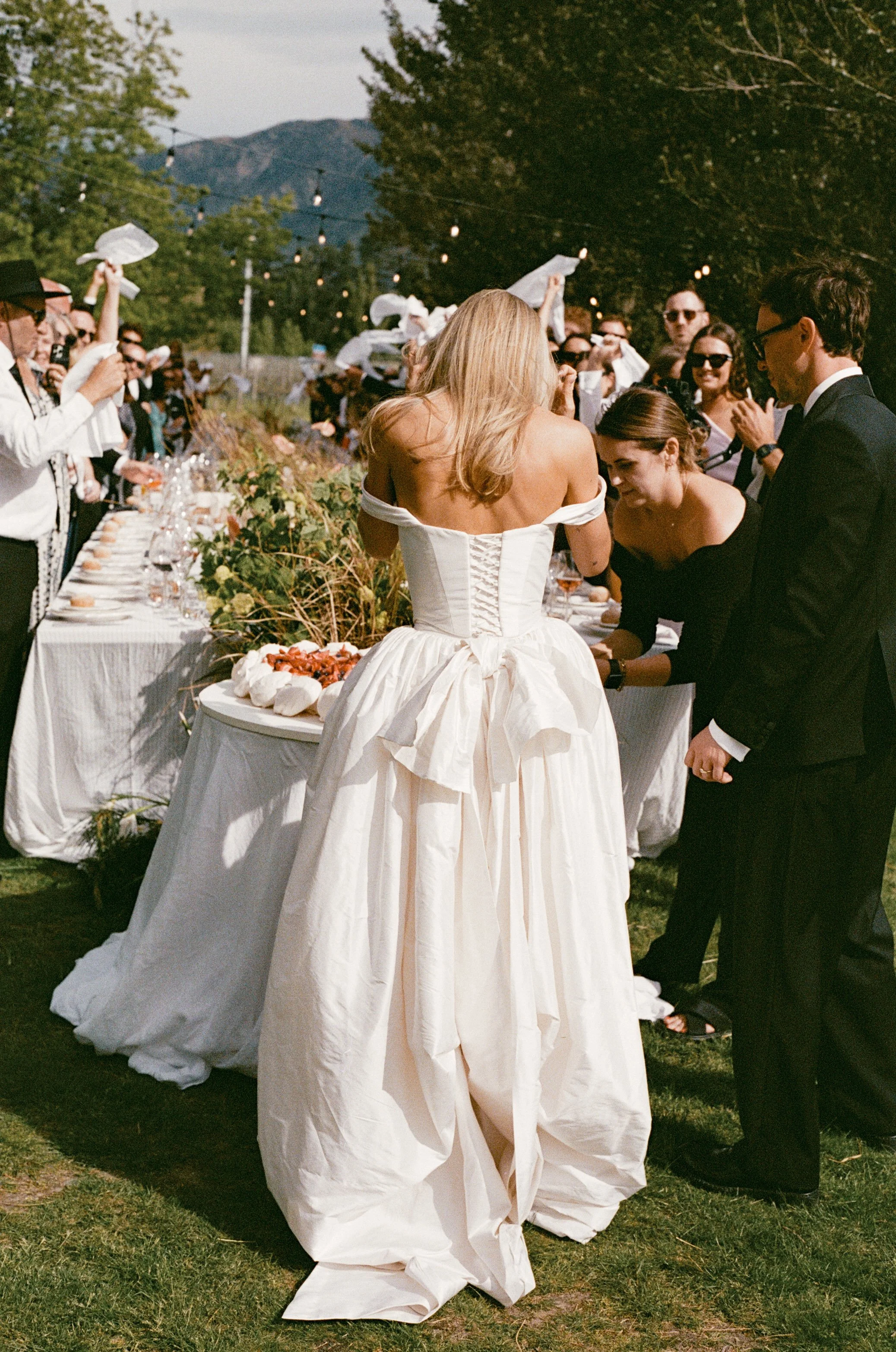 Wedding reception outdoors with a long table set with flowers, food, glasses, and white linens, surrounded by guests in formal attire, greenery, trees, hanging lights, and mountains in the background.