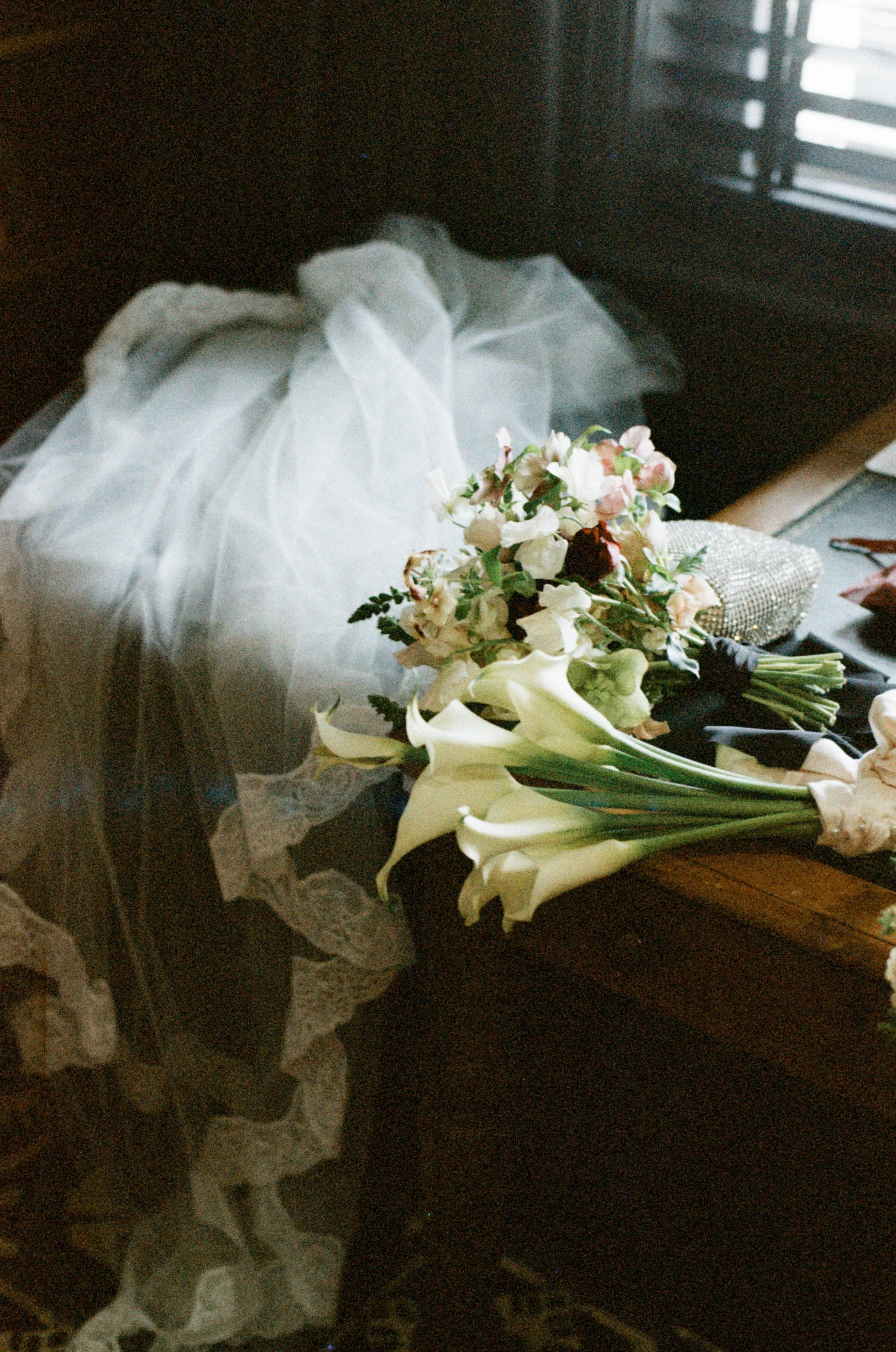 A floral bouquet of white and pink flowers resting on a dark wooden surface near a window with blinds, with a white dress or tulle fabric draped nearby.