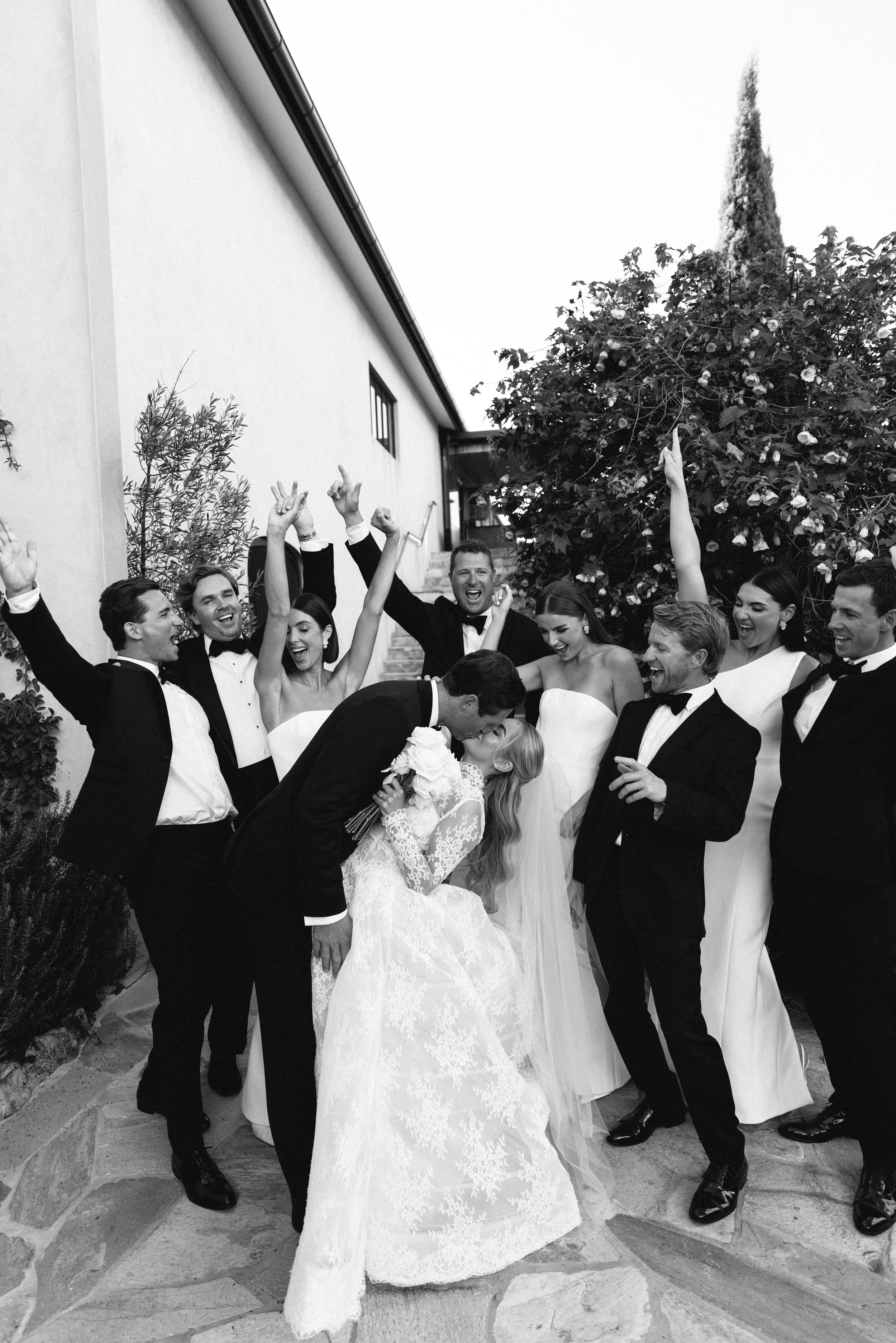 Black and white photograph of a bride and groom surrounded by wedding guests, celebratorily raising their hands outdoors with a building, bushes, and a tall tree in the background.
