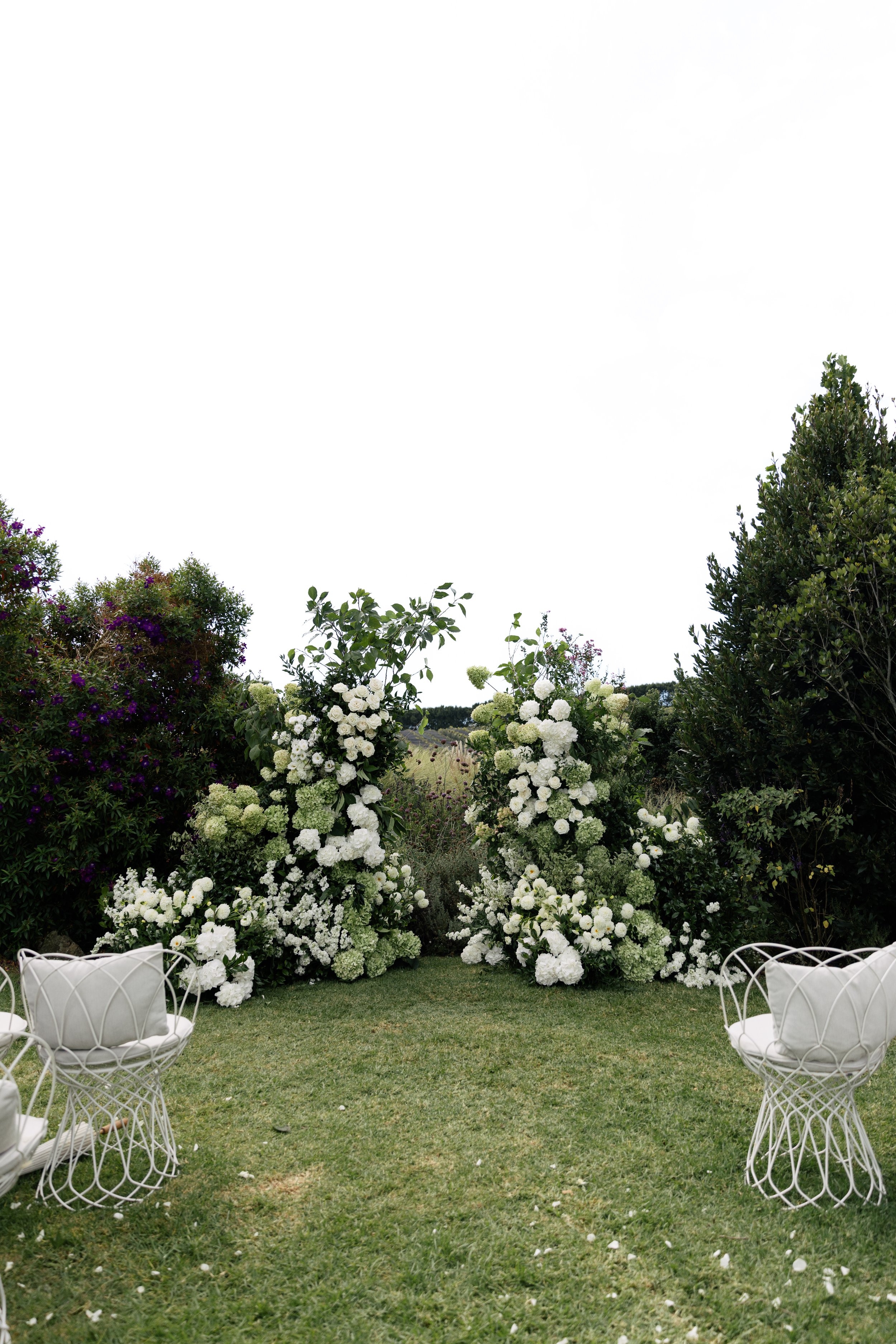 Outdoor garden wedding setup with white chairs, lush green grass, and a floral backdrop of white and purple flowers with green foliage.