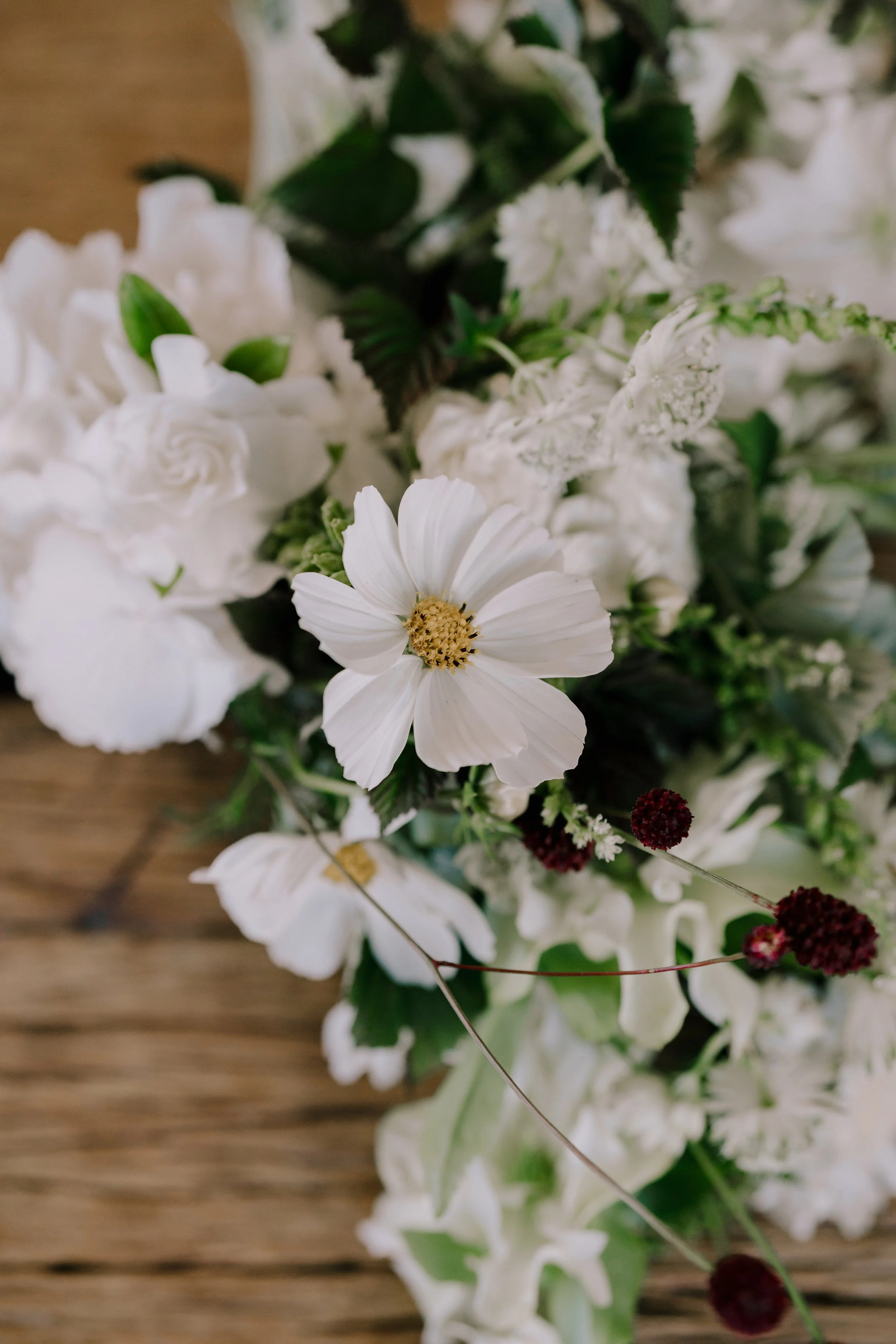Close-up of a floral arrangement with white flowers, green leaves, and small dark red berries on a wooden surface.