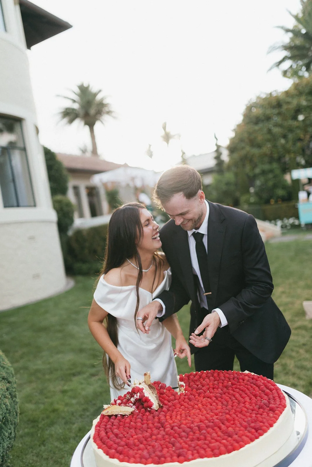 A man and woman laughing and smiling at a wedding cake outdoors, woman in a white dress and man in a black suit, with a large round cake topped with raspberries in front of them.
