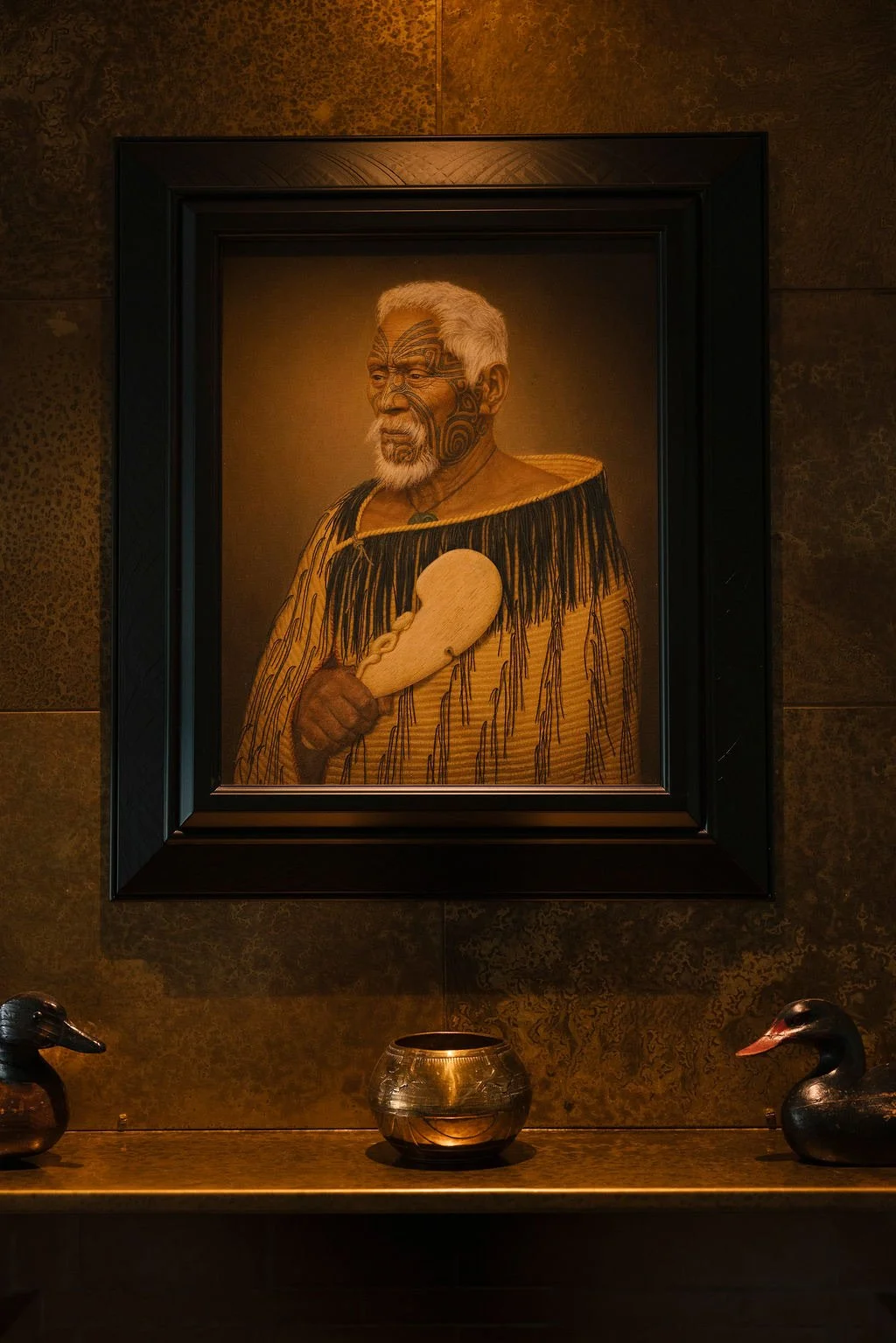 Portrait of an elder Polynesian man with tribal tattoos, holding a wooden paddle, dressed in traditional attire with fringe details, framed on a wall with decorative items below.