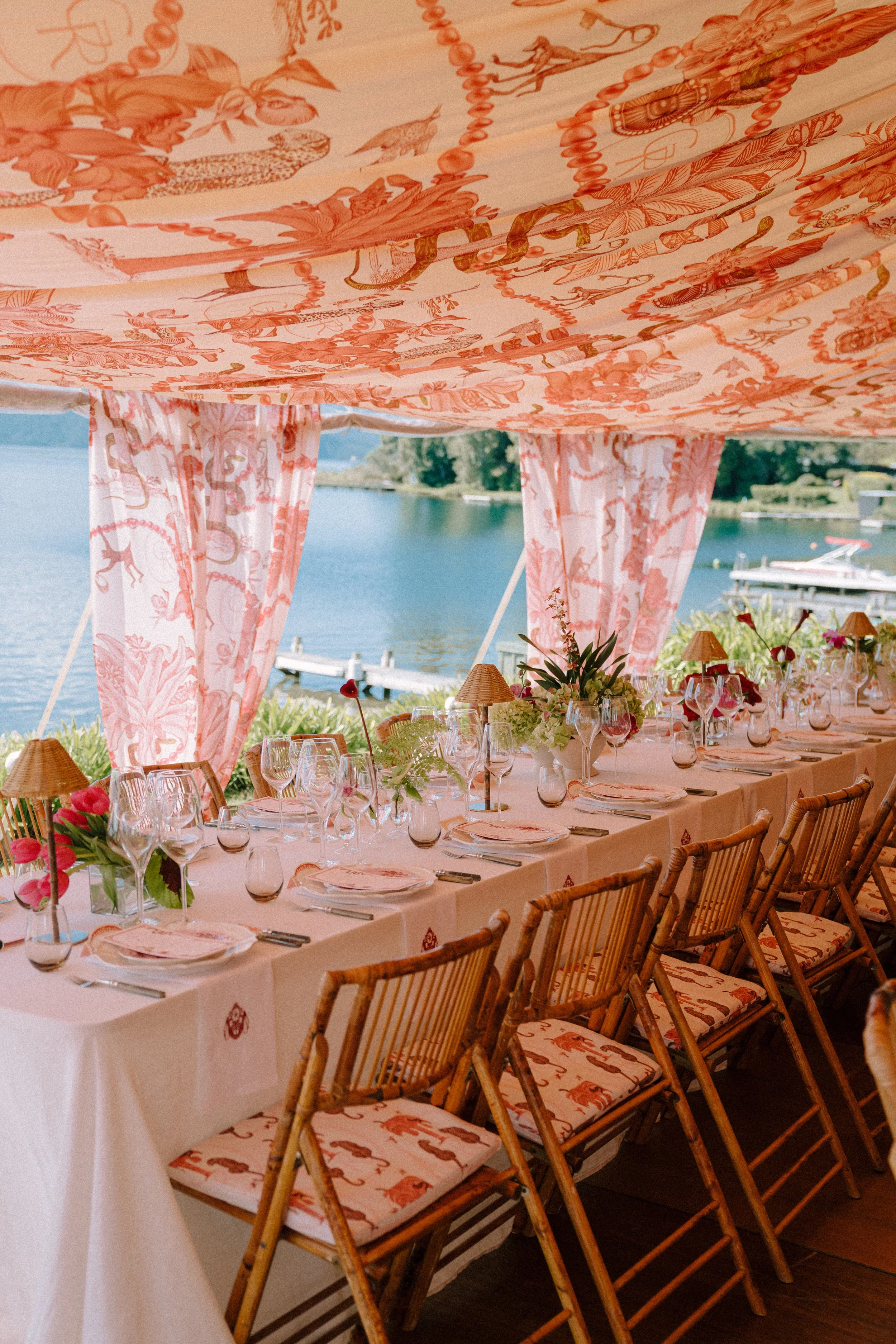 A long banquet table set for a meal outdoors near a body of water, under a pink and orange canopy decorated with animal and floral patterns, with chairs around the table.