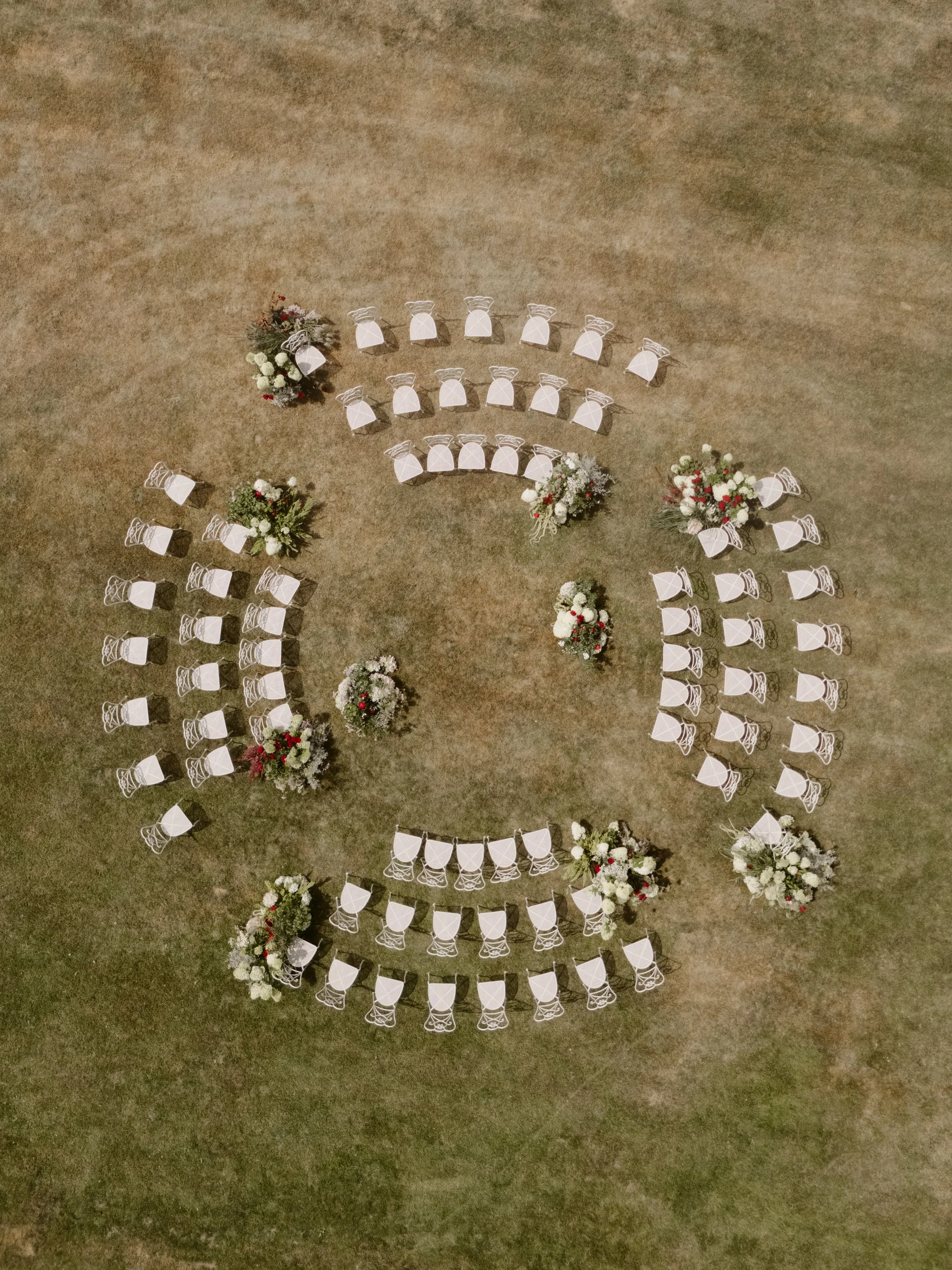 Empty white chairs arranged in rows with floral arrangements at the ends, set up on a dirt ground for an outdoor event, likely a wedding ceremony.