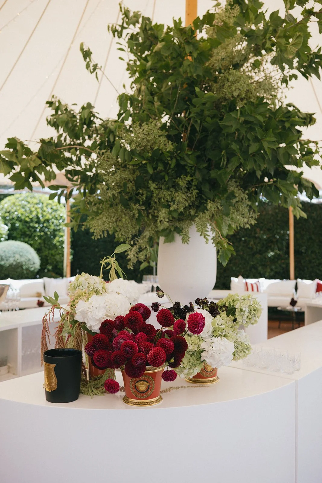 Large white vase with green leafy plants and white flowers, surrounded by smaller vases with red berries and white flowers, on a white curved surface, outdoors with greenery in the background.