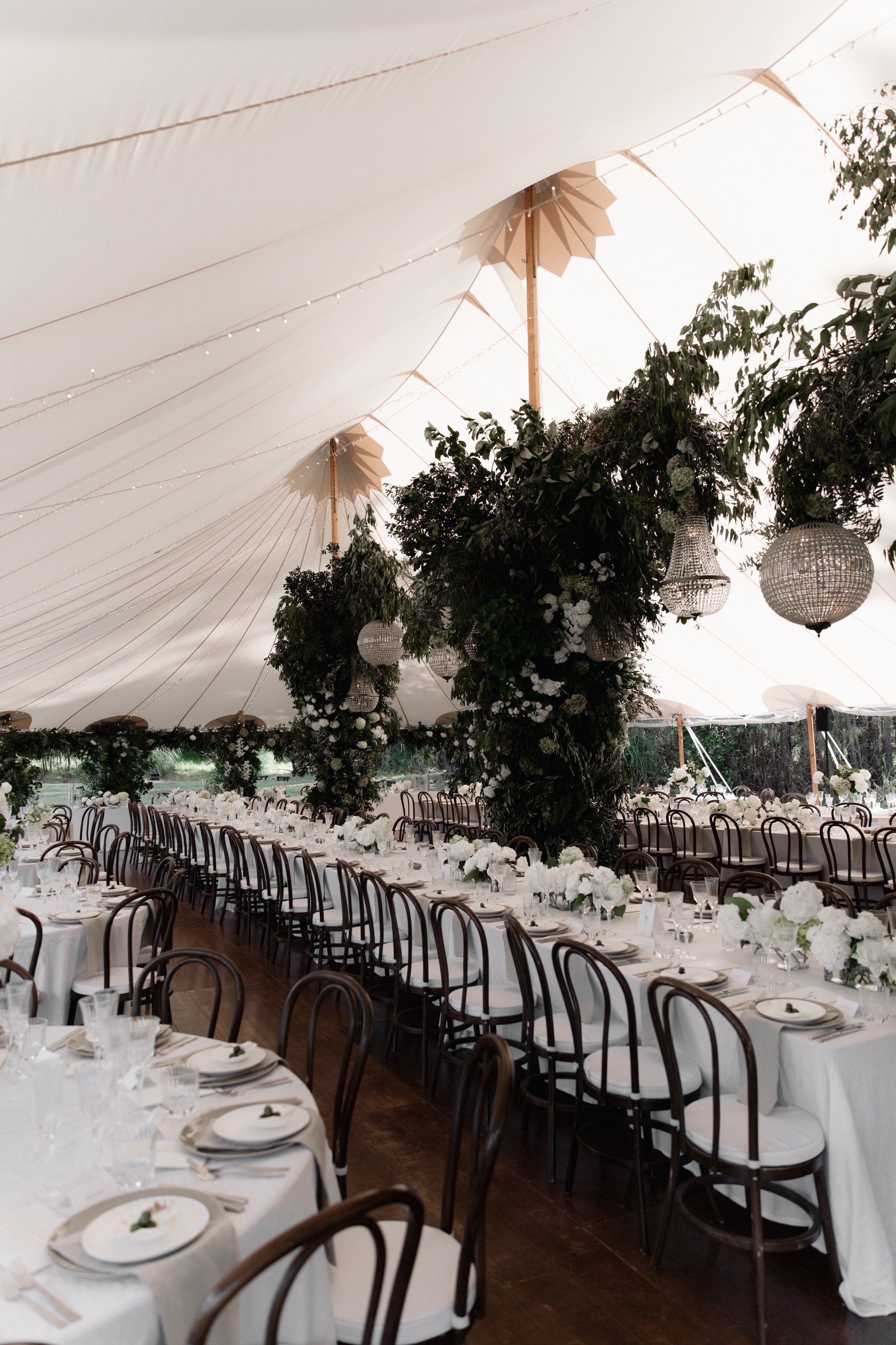 Elegant banquet hall decorated with white flowers, greenery, and crystal chandeliers under a large white tent.