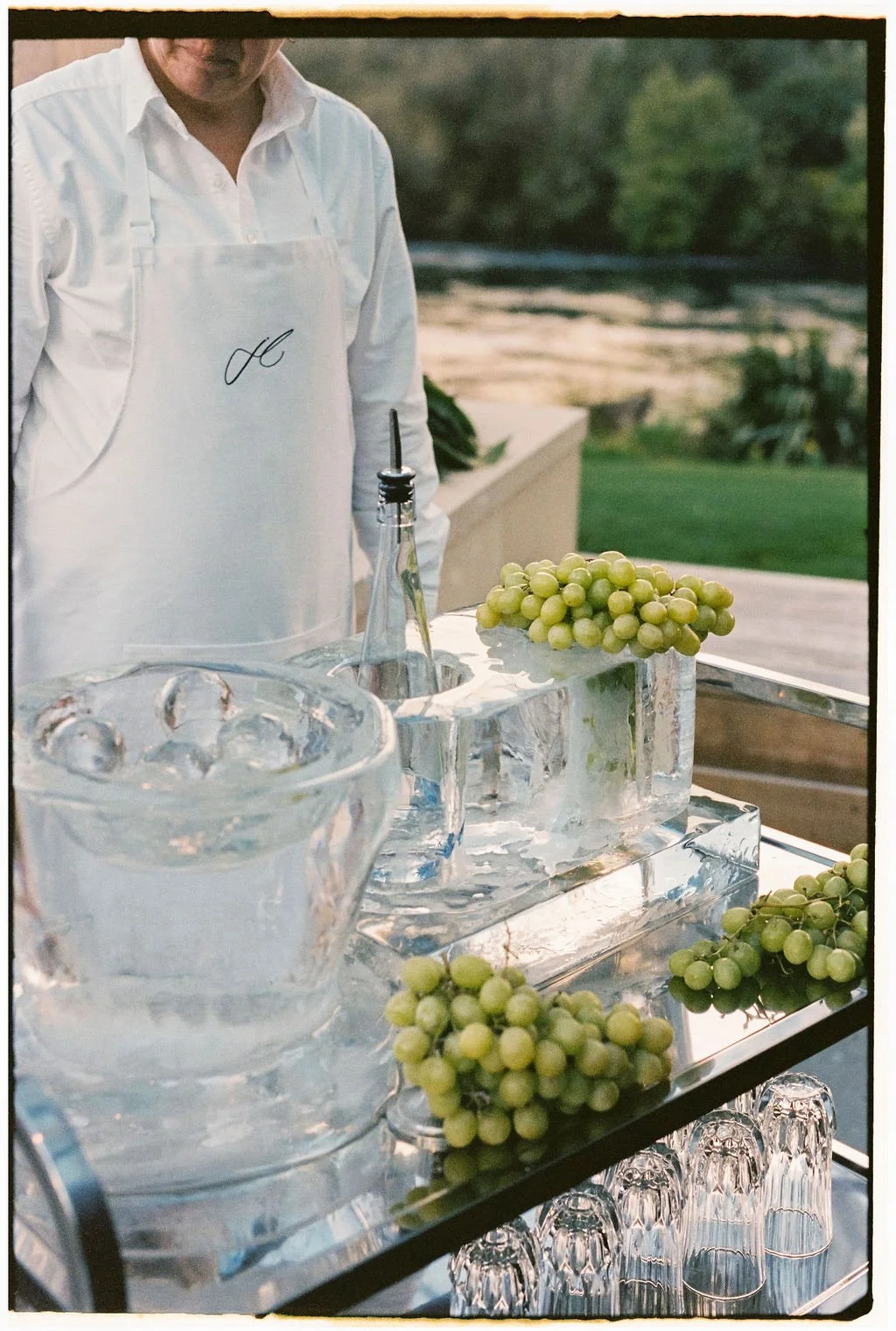 Ice bucket with grapes and a bottle of wine or champagne on a glass table outdoors, with a person in a white shirt and apron behind, and a grassy yard in the background.
