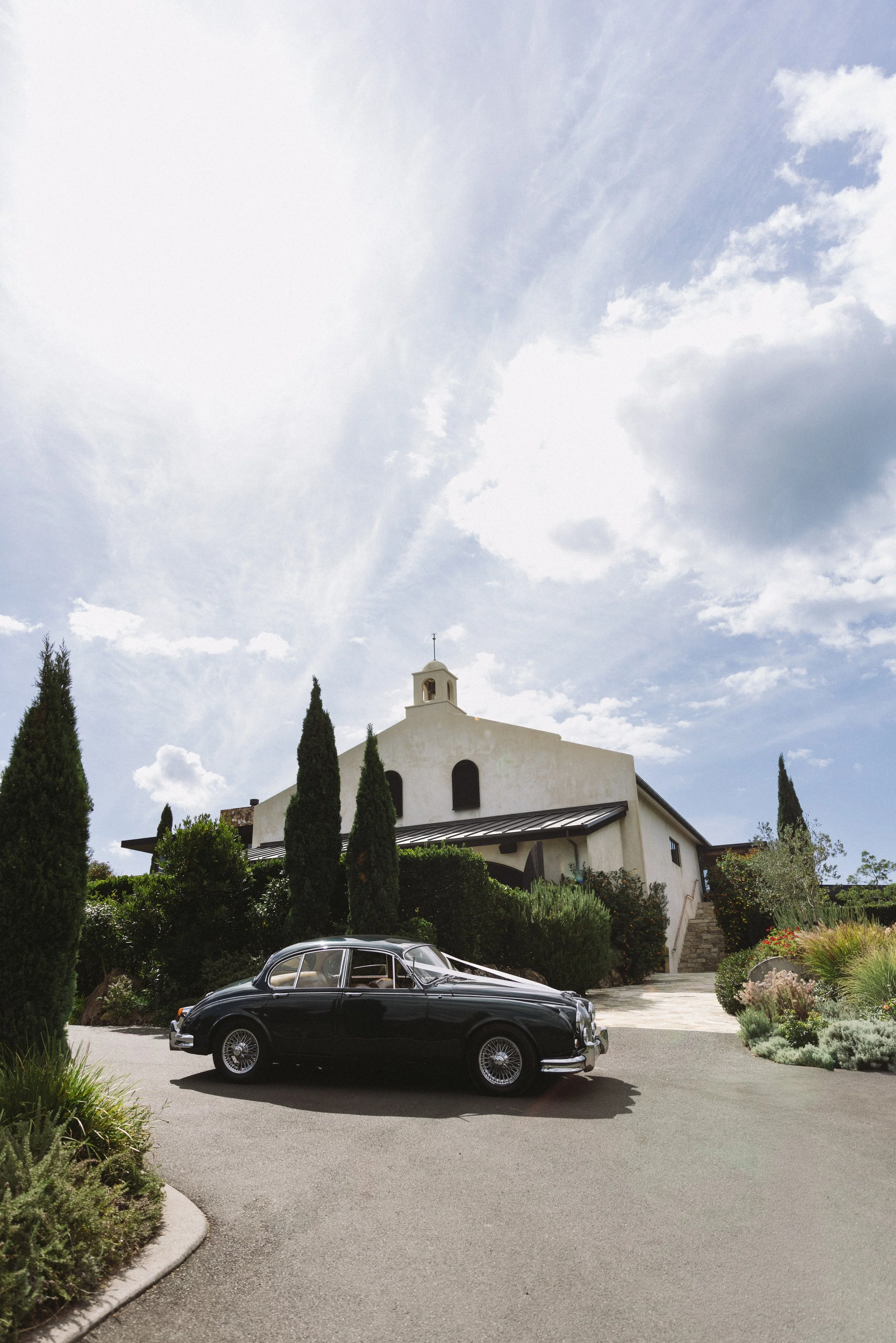 A black vintage car parked in front of a white building with a bell tower. The building is surrounded by tall, narrow cypress trees and lush greenery. The sky is partly cloudy.