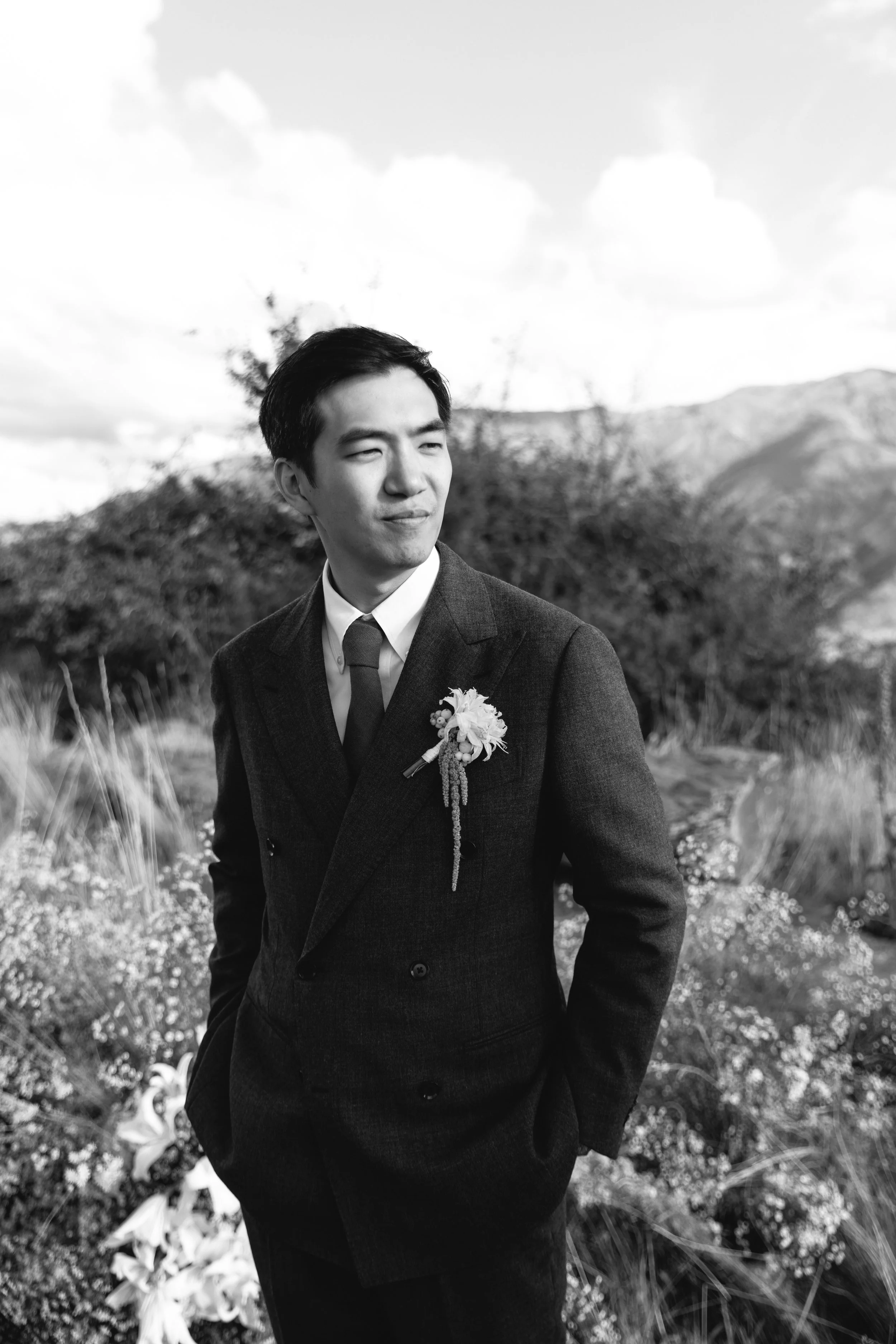 Black and white photo of a man in a suit standing outdoors in a natural setting with mountains in the background and a cloudy sky.