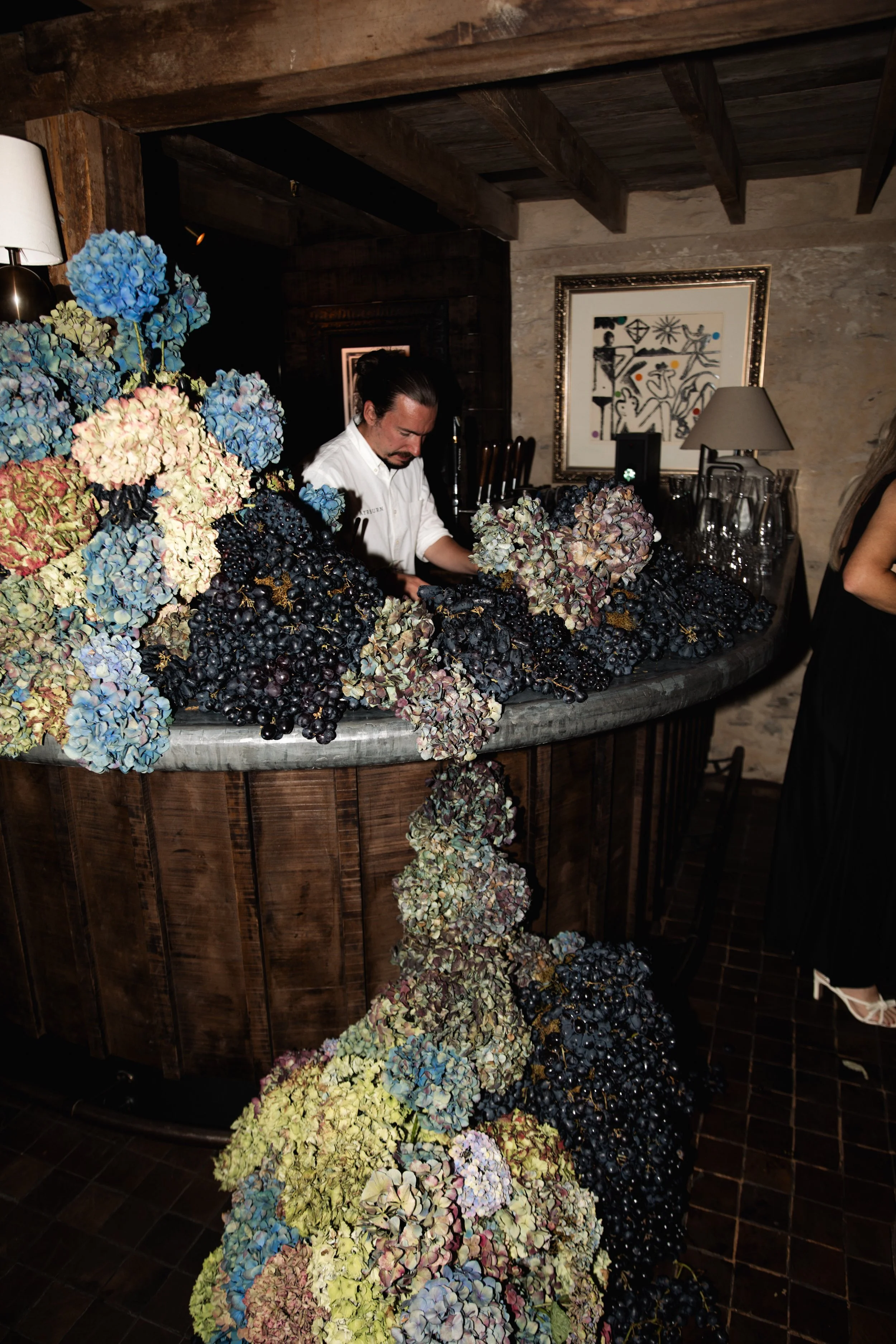 An individual standing behind a bar surrounded by large clusters of colorful hydrangea flowers and dark grapes, in a rustic indoor setting.