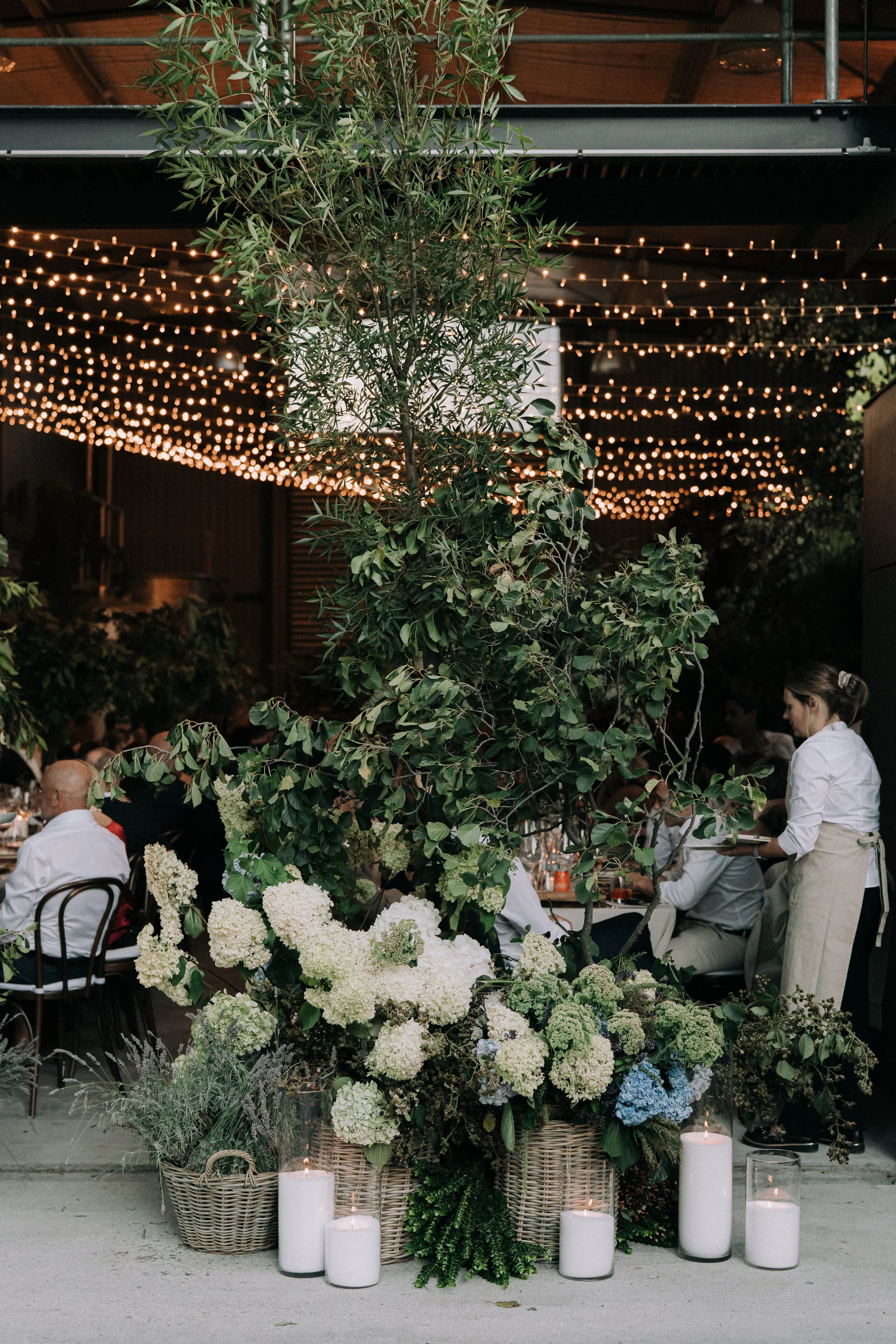 Elegant floral centerpiece with white hydrangeas, green foliage, and candles in woven baskets set on a table at a wedding reception with string lights overhead.