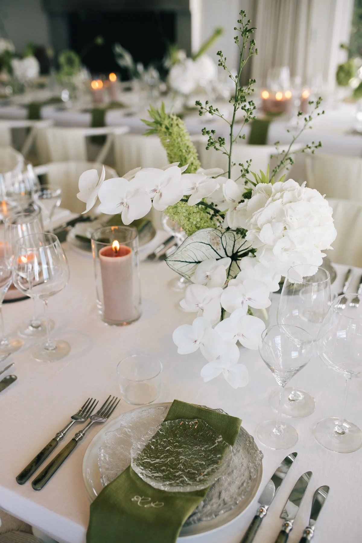 Close-up of an elegantly set dining table with a floral centerpiece of white orchids, hydrangeas, and greenery, with candles and glassware in the background.