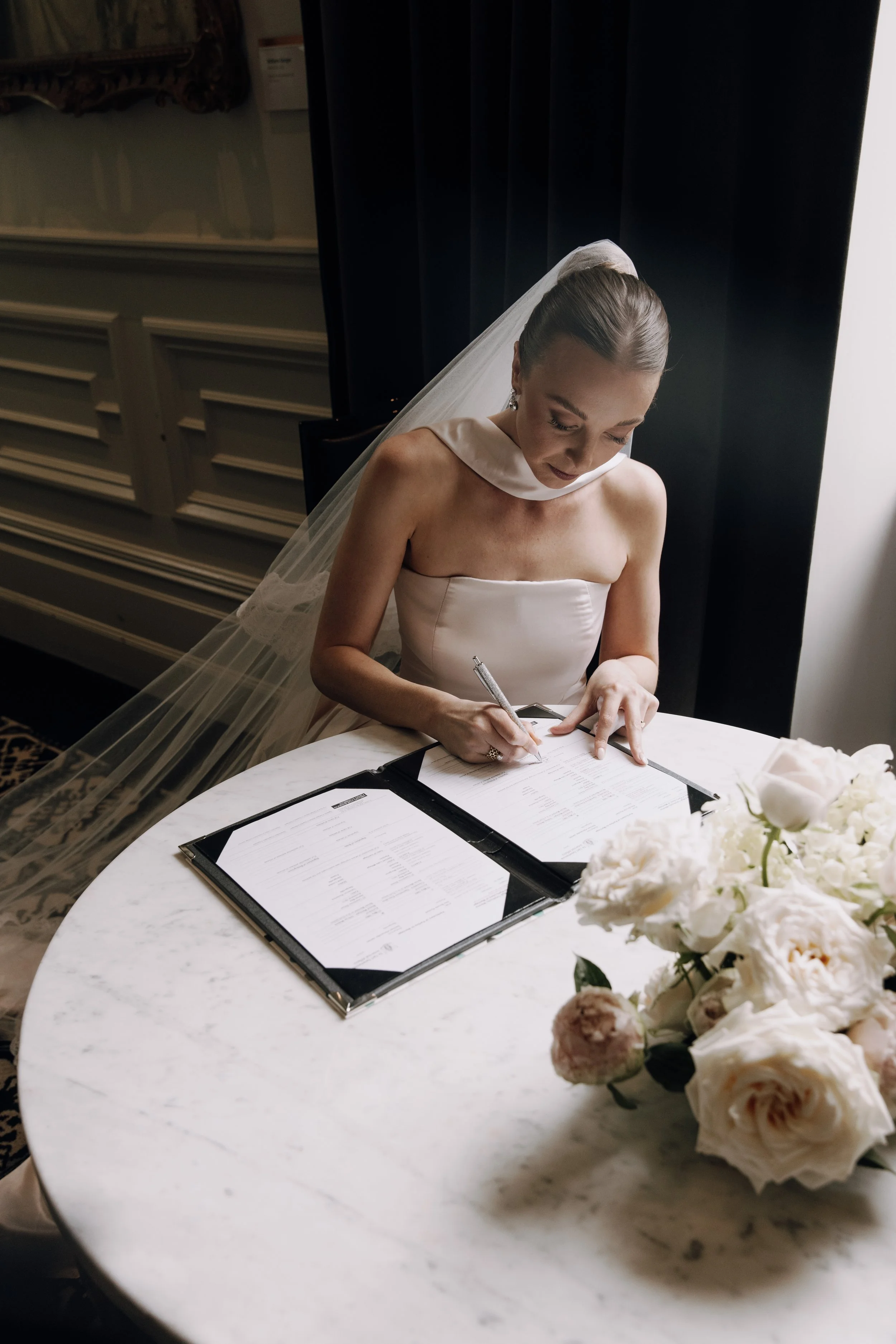 A bride in a white strapless wedding gown with a satin satin collar and matching veil signing a document at a round marble table, with a bouquet of white and pink flowers nearby.