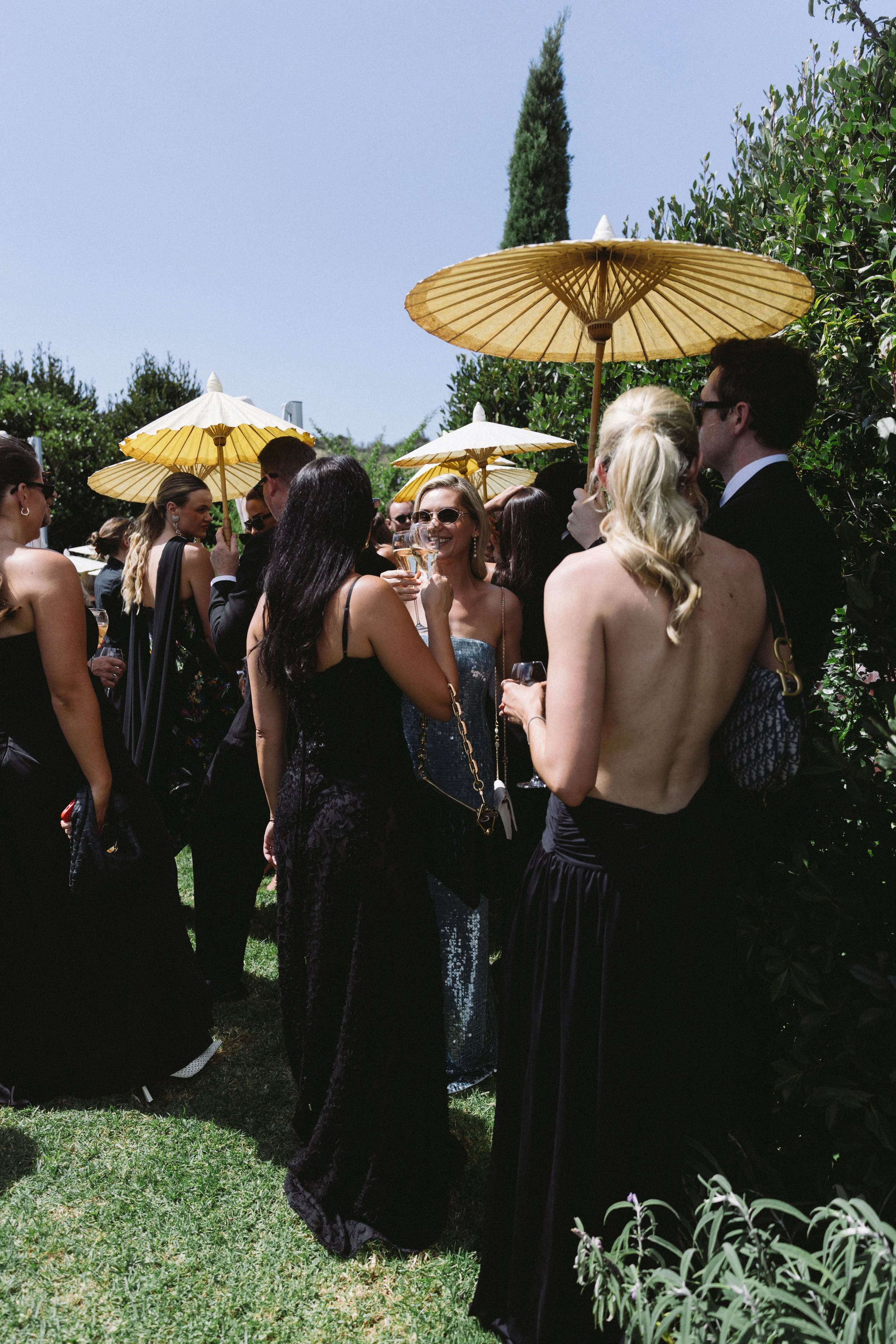 People dressed in formal attire at an outdoor event, holding drinks, with yellow umbrellas and lush greenery under a clear sky.