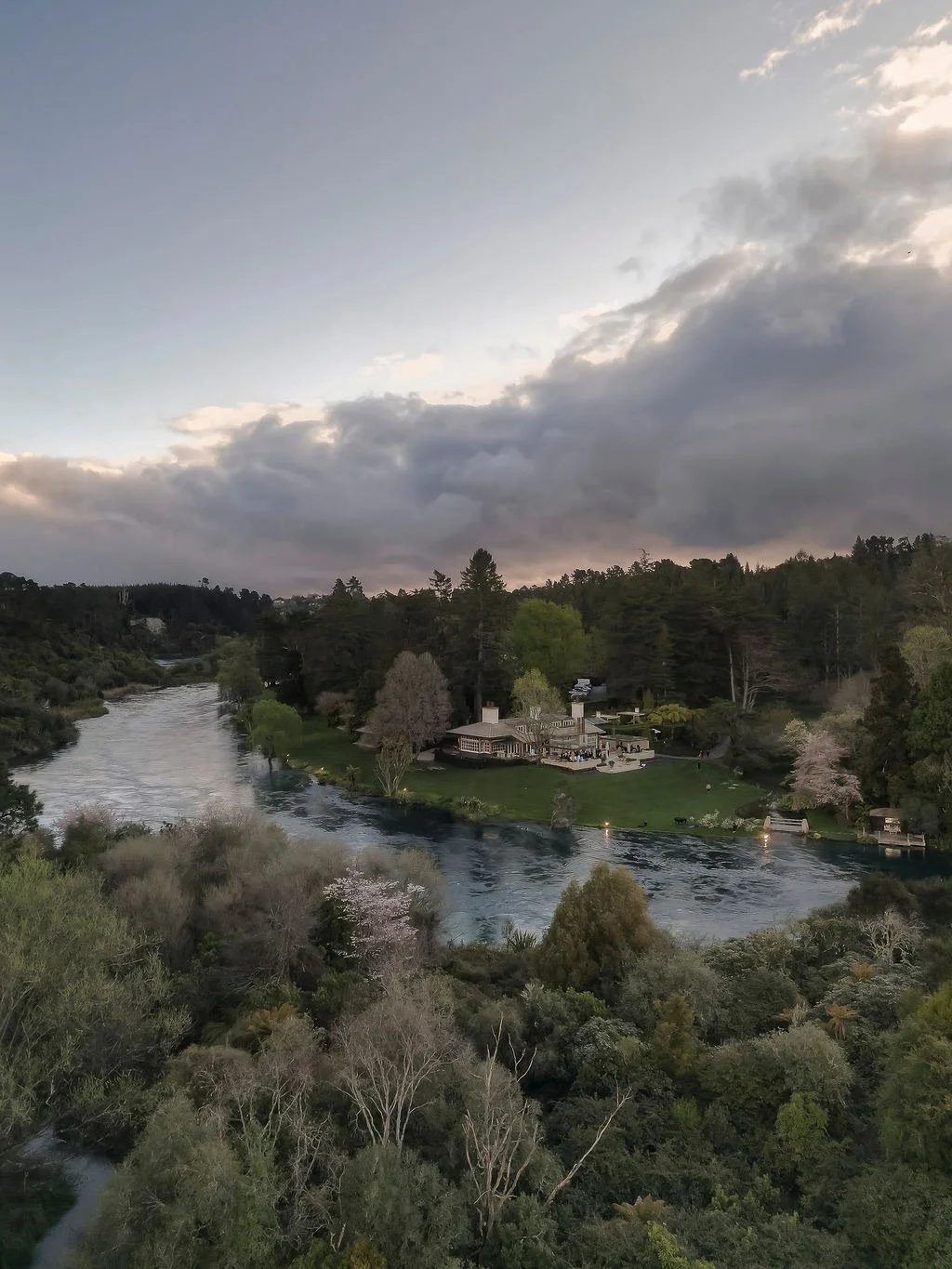 A scenic aerial view of a river flowing through a lush green landscape with a house on the riverbank, surrounded by trees, with a cloudy sky overhead.
