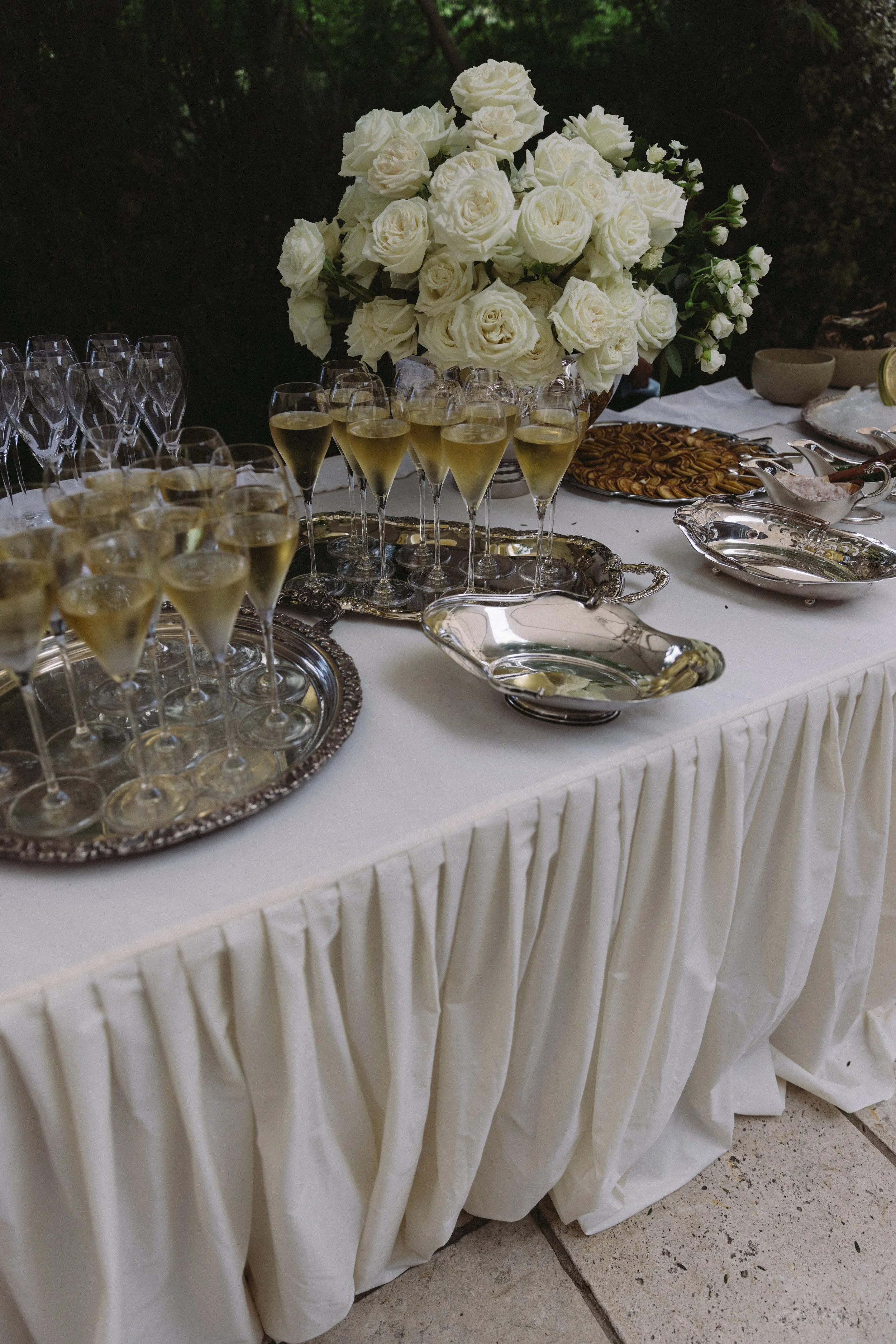 A table with a white tablecloth decorated with pleated fabric skirting, holding a large bouquet of white roses in a vase, several glasses of champagne, and a platter of pecans. There are additional serving dishes and bowls on the table, with a dark, outdoor background.