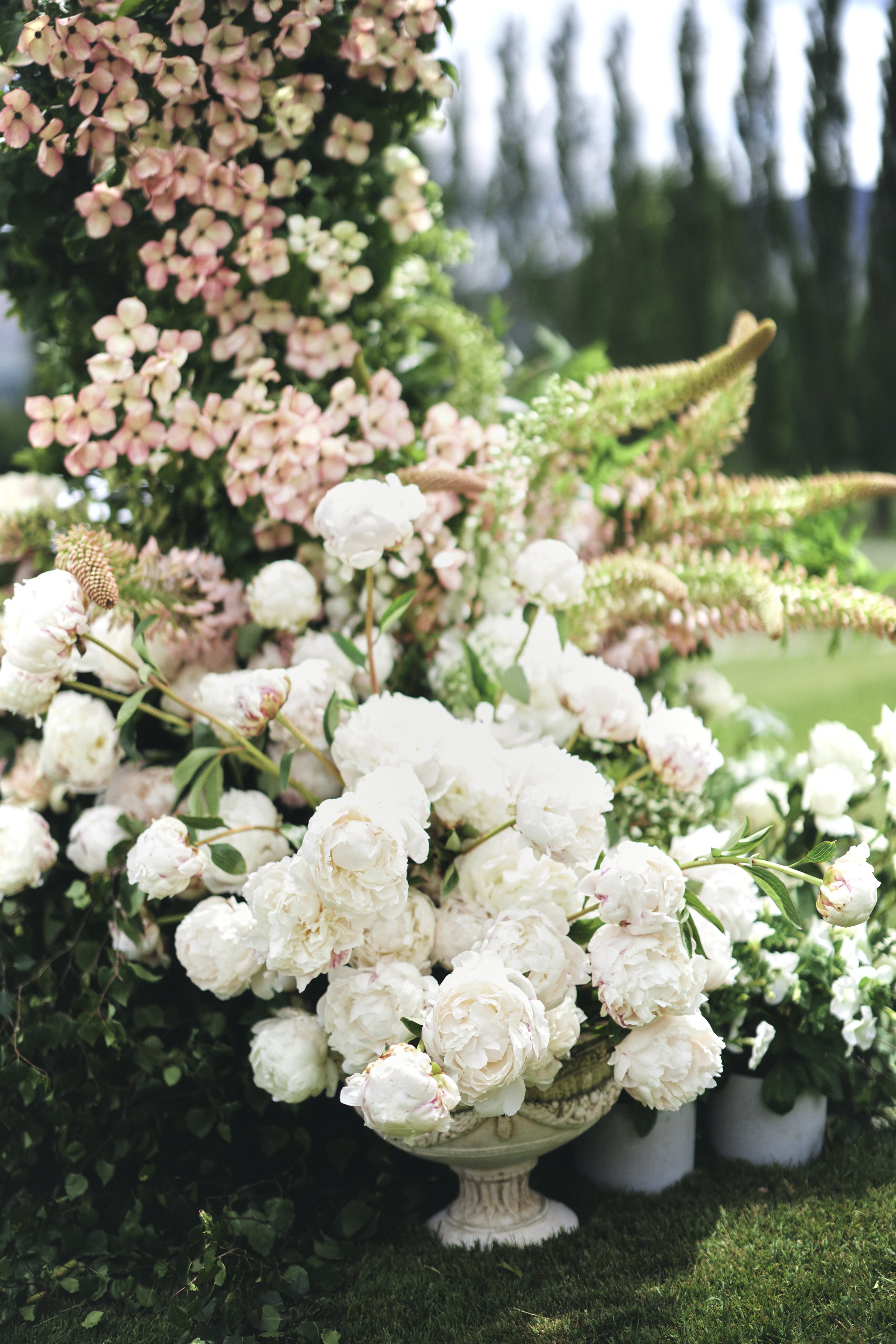 White peonies arranged in a decorative urn outdoors, with pink and white hydrangeas and greenery in the background.
