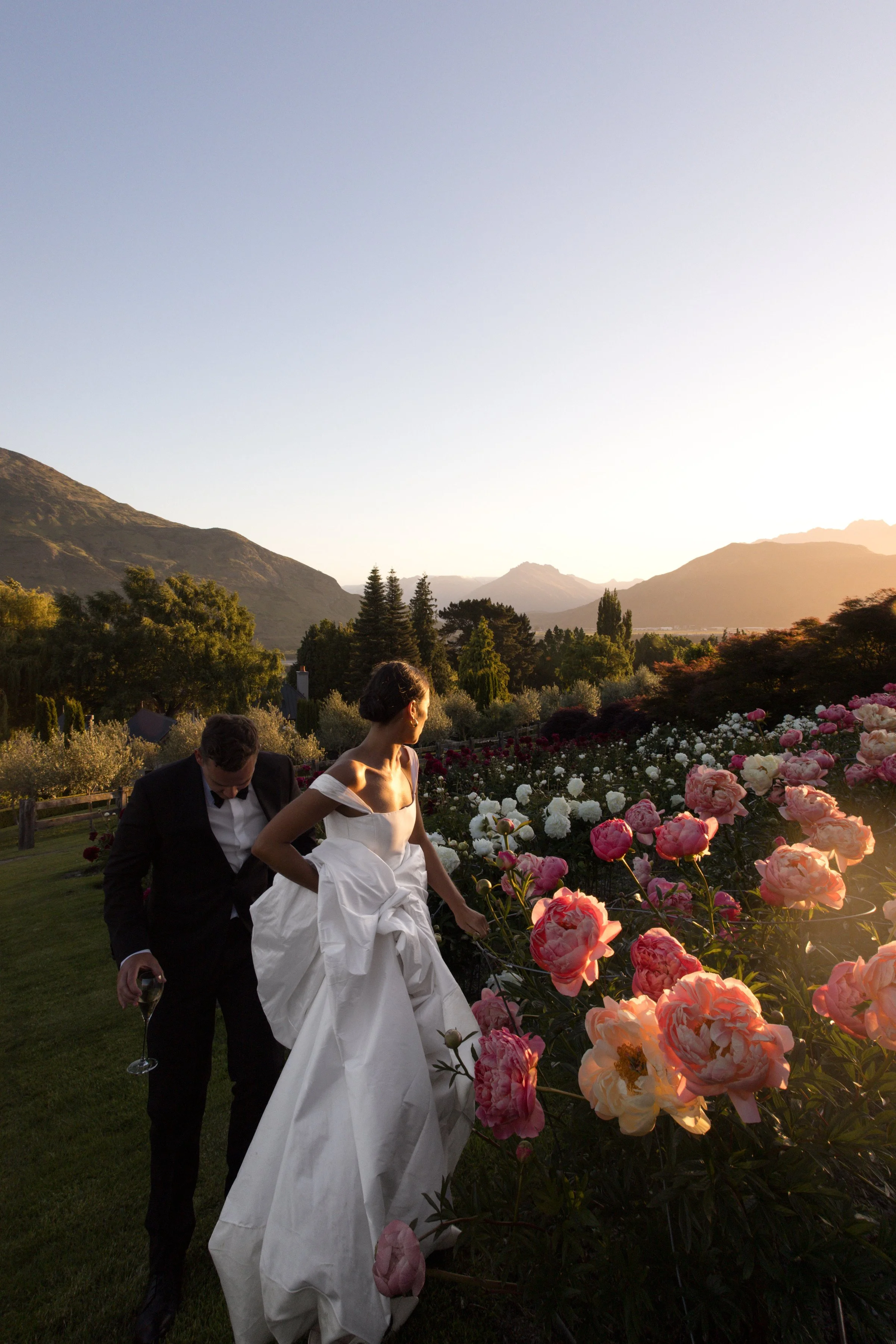 A bride and groom walking through a garden of pink and white flowers during sunset with mountains in the background.