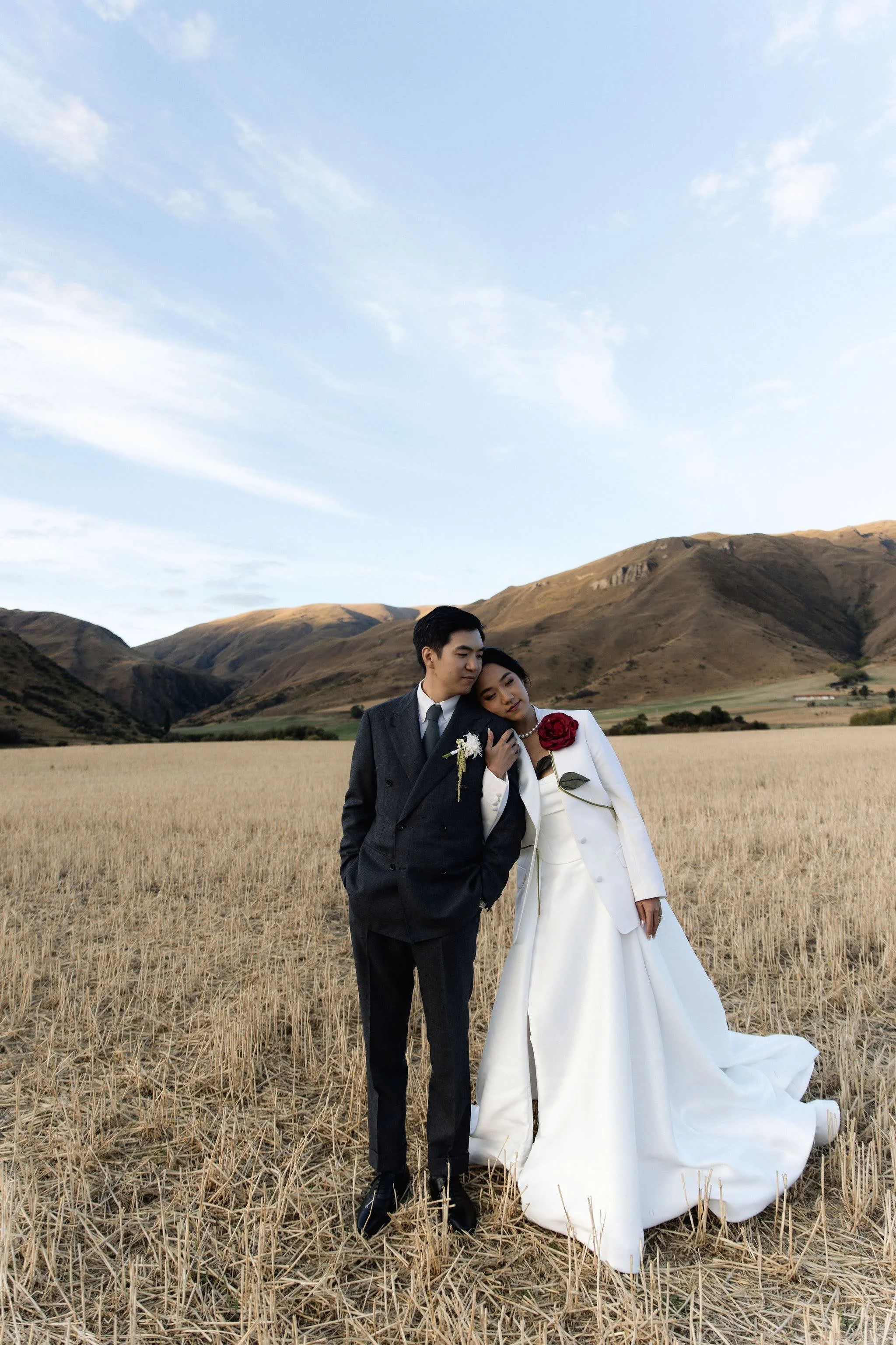 A bride and groom standing in a field of dried grass with rolling hills in the background. The groom is wearing a dark suit and the bride is in a white wedding dress holding a red rose. The woman leans her head on the man's shoulder.