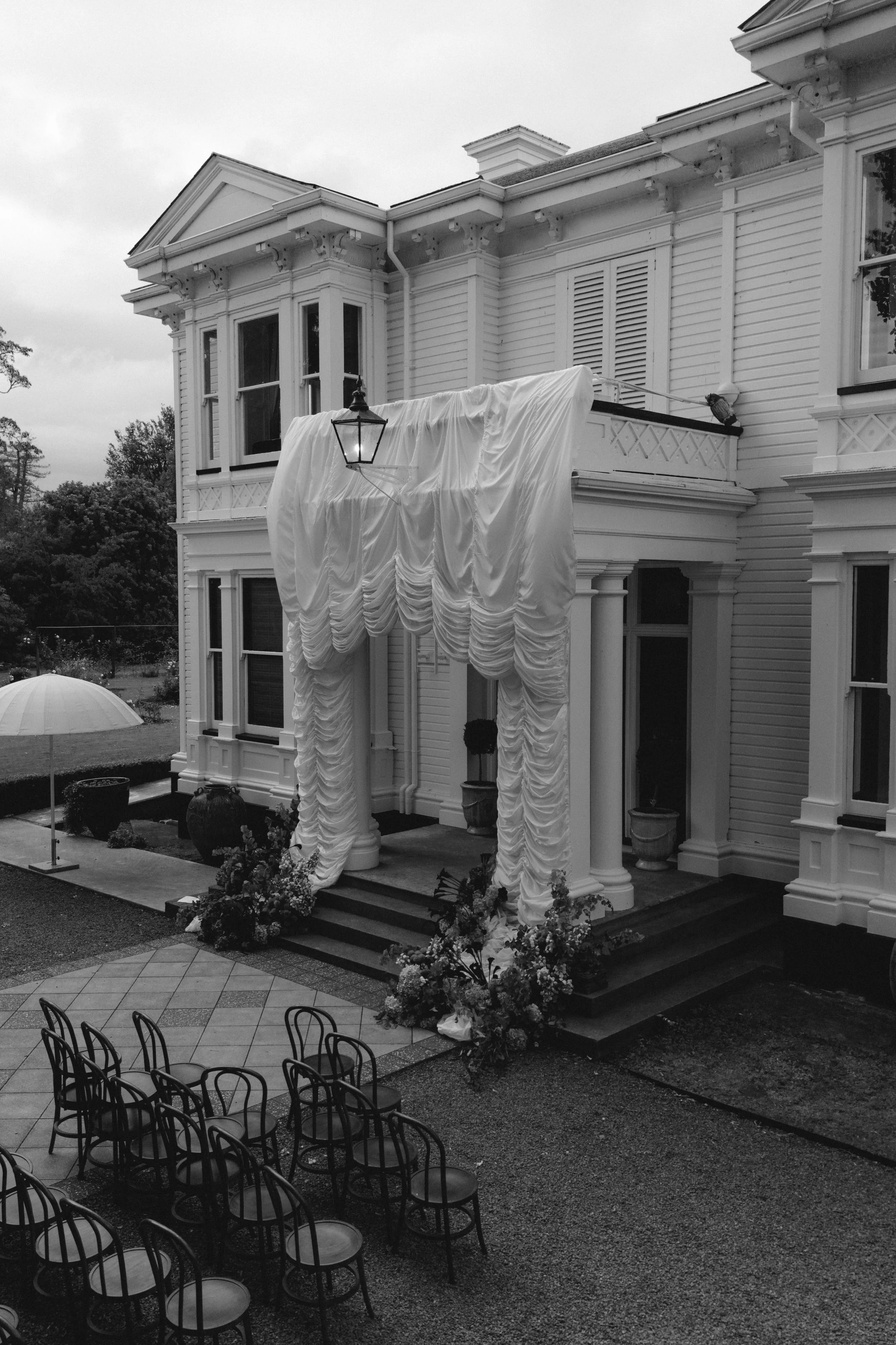 Black and white photo of a large house with classic architectural details, set up for a wedding or event with draped fabric, flowers, and chairs arranged outdoors.