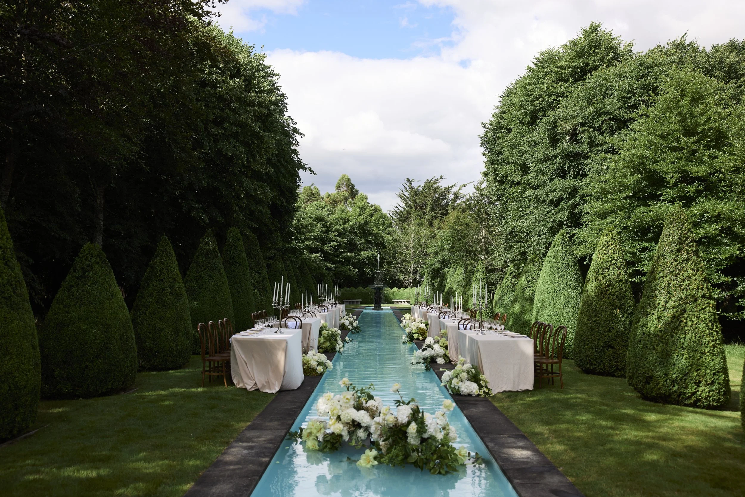 Outdoor wedding ceremony setup with tables on both sides of a water aisle, surrounded by tall green trees and manicured bushes, under partly cloudy sky.